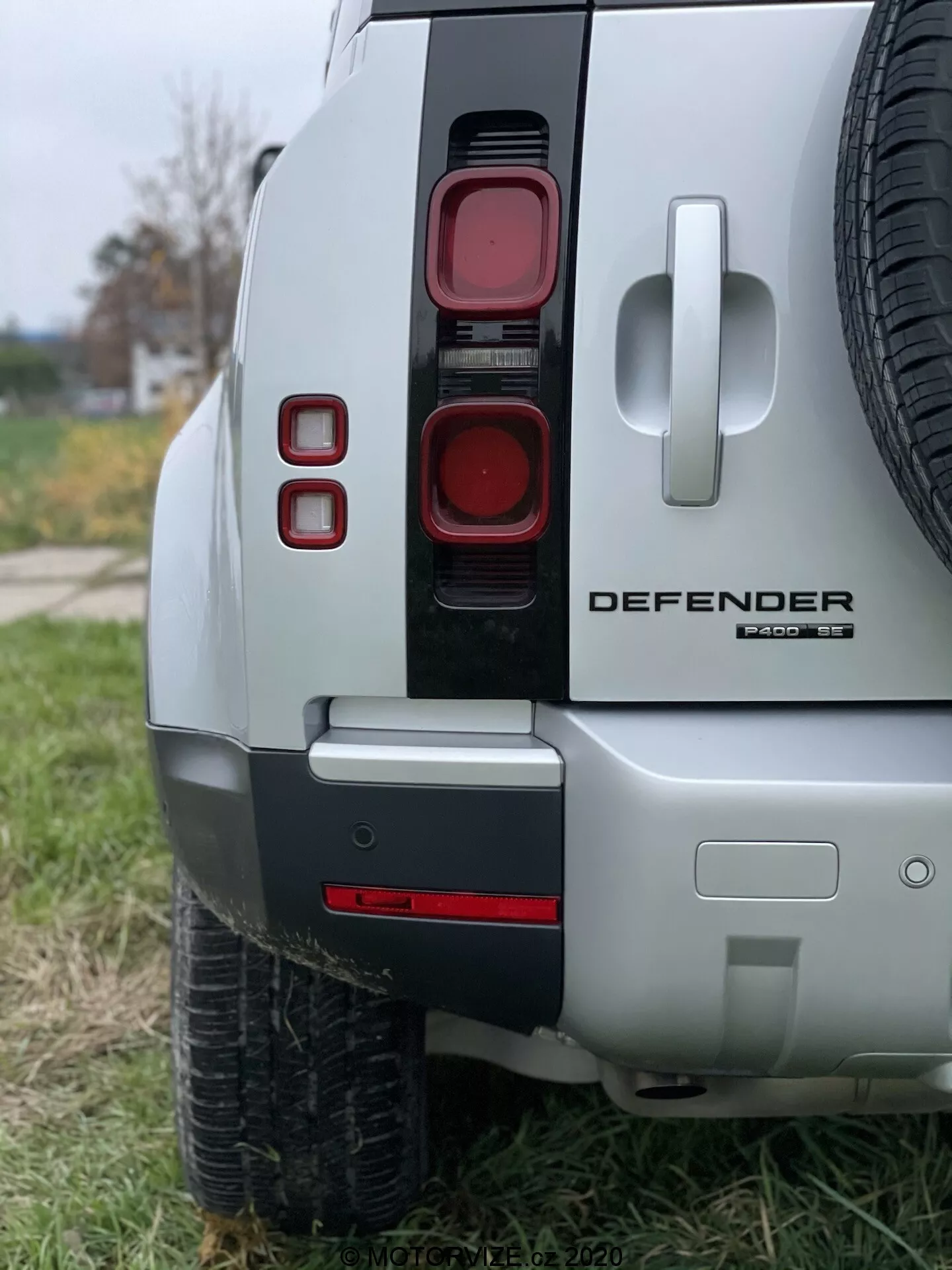 Bližnji posnetek zadnjega dela Land Rover Defender 2020 prikazuje sodobne navpično razvrščene zadnje luči, izraziti oznaki 'DEFENDER' in 'X-Dynamic SE', delni pogled na rezervno pnevmatiko na zadnjih vratih, dvo-barvni odbijač z morebitno vlečno kljuko in srebrno barvo karoserije, kar poudarja prefinjen in terenski dizajn vozila.