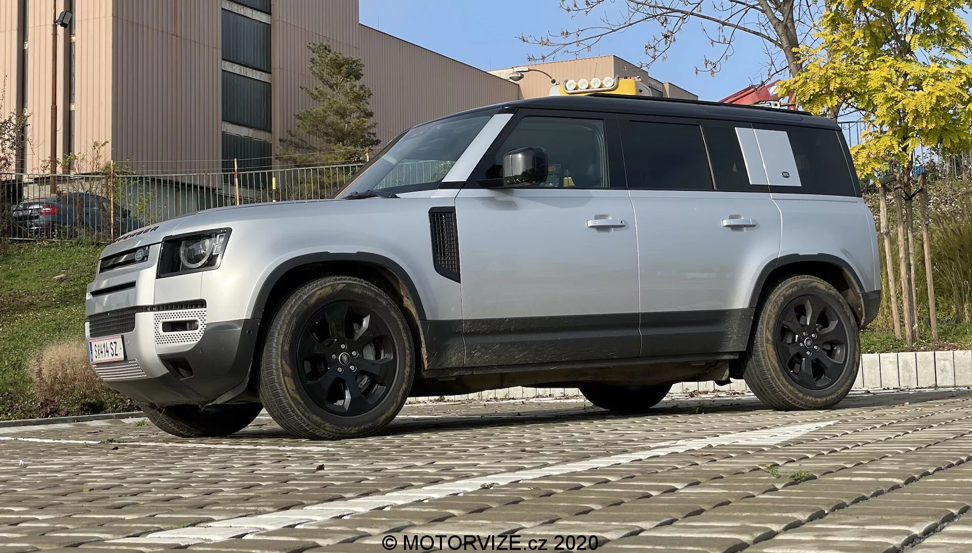 Fotografija modernega SUV-ja Land Rover Defender 2020, posneta iz nizkega kota, da poudari njegovo robustno in impozantno držo. Vozilo je prikazano v trideset četrtinskem pogledu s sprednje in bočne strani, kar poudarja njegovo značilno mrežo, LED žaromete, odbijač z meglenkami in senzorji, črne aluminijaste platišča z večprečnim dizajnom in nizkoprofilnimi pnevmatikami, črna bočna ogledala s potencialnimi dodatnimi tehnologijami, zatemnjena okna, izjemno linijo strehe, izplaknjene ročaje vrat, močan dizajn kolesnih lokov in svetlo srebrno ali sivo barvo, ki kontrastira s črnimi elementi. SUV je parkiran na tlakovani podlagi, s stavbo in listjem v ozadju pri dnevni svetlobi.