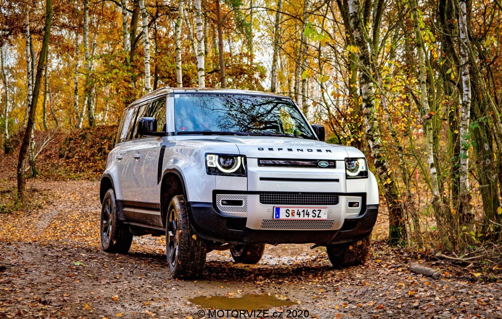Ta slika prikazuje SUV Land Rover Defender 2020, ki je postavljen na neurejeni gozdni poti, prikazuje sodoben, oglat dizajn iz sprednjega tridimenzionalnega kota. Jasno vidni so rešetka, žarometi in bočni profil vozila. Osvetljeni so značilni LED žarometi z dnevnimi lučmi, izrazita rešetka in poudarjeni meglenki. Velika aluminijasta kolesa z off-road tekalno plastjo, stranska ogledala z vgrajenimi smerniki ter dvignjena oddaljenost od tal poudarjajo njegove off-road sposobnosti. Robusten videz, tradicionalni ročaji vrat in nevtralna barva karoserije še povečajo njegov privlačni status kot sodoben SUV, pripravljen na avanture.