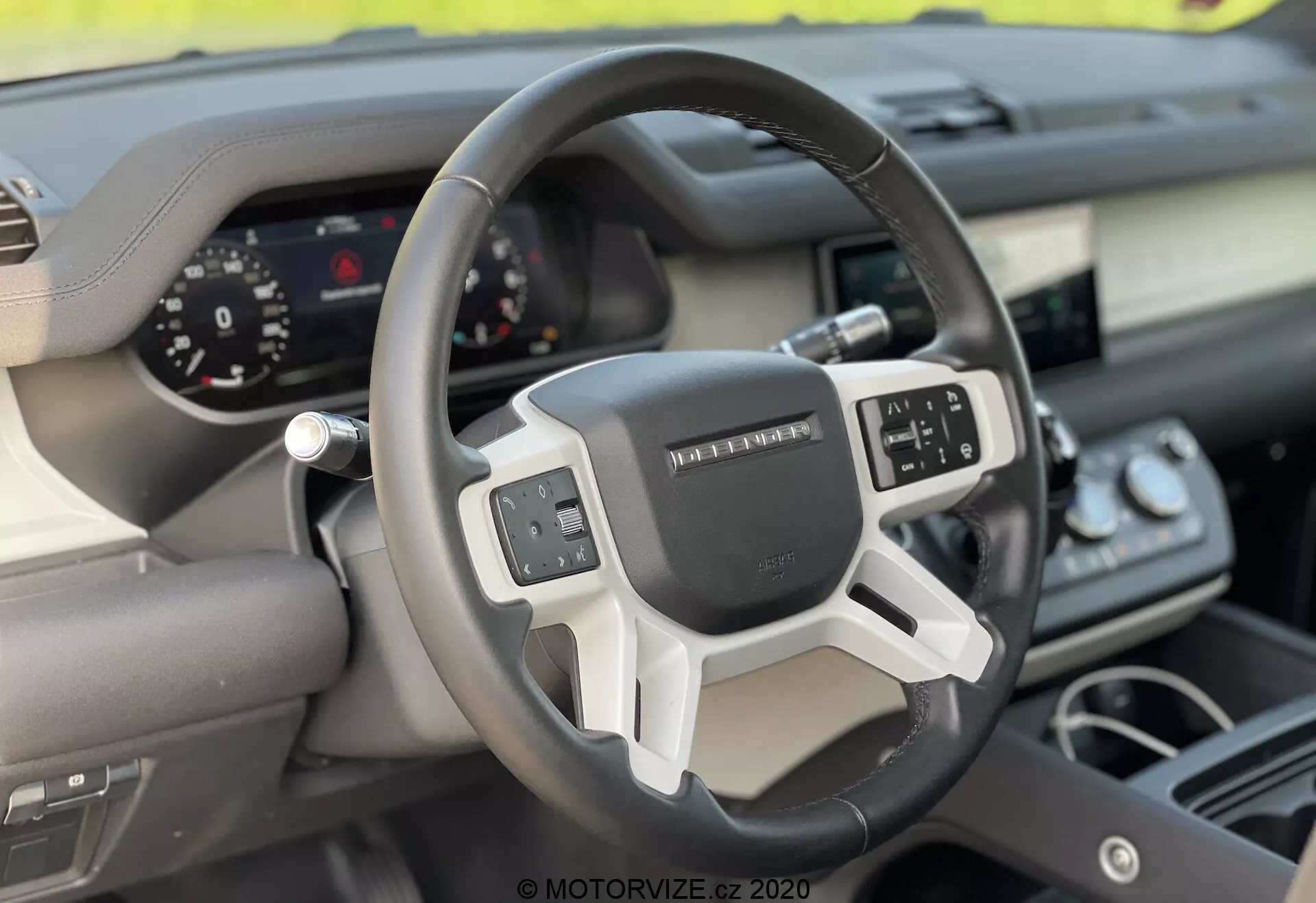 Pogled v notranjost Land Rover Defender 2020, osredotočen na volan in armaturno ploščo z vidika prednjega sovoznikovega sedeža. Moderni trikraki volan ima večfunkcijske kontrole in digitalni instrumentni grozd za jasen prikaz merilnikov. Mešanica materialov, kot so usnje in kovinski ali plastični dodatki, poudarja ergonomsko in tehnološko zasnovo vozila.