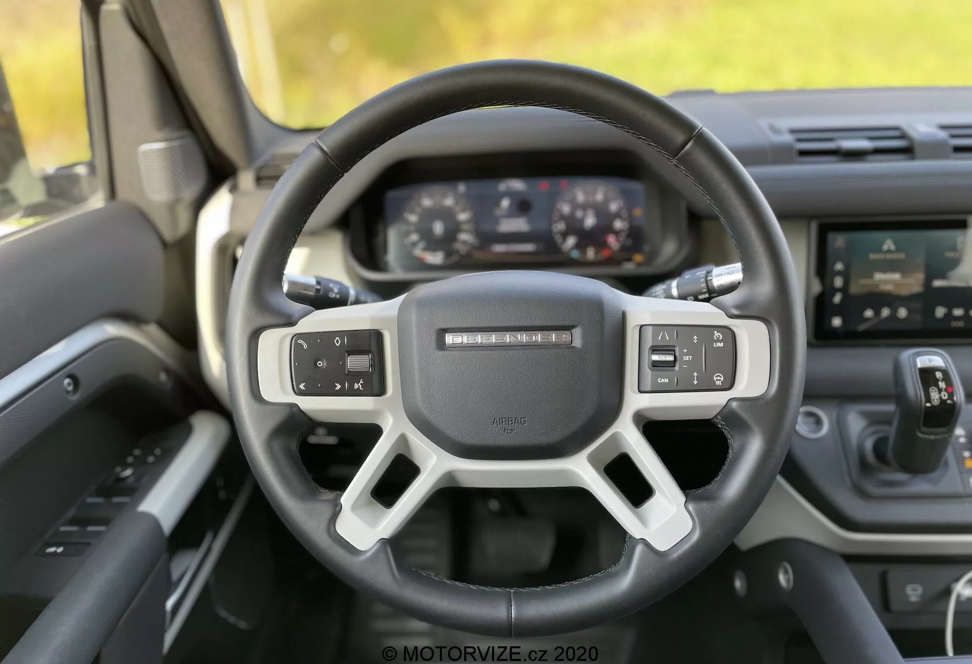 Notranji pogled Land Rover Defender 2020, prikazano iz perspektive voznika s poudarkom na volanu in armaturni plošči. Trokrajni volan z ravnim dnom ima večfunkcijske kontrole in zbluriran znak v sredini. Za njim je digitalen instrumentni panel, ki prikazuje hitrost in druge informacije, kar kaže na moderno tehnologijo. Sredinska konzola prikazuje sistem za informacije in zabavo na dotik s različnimi ikonami in fizičnimi gumbi za nadzor klimatskih in avdio funkcij. Notranjost je izdelana iz trajnega materiala, verjetno iz trde plastike ali ogljikovih vlaken. Visokokakovostna slika, posneta pri naravni svetlobi, uporablja bokeh učinek za poudarjanje volana in kontrol.