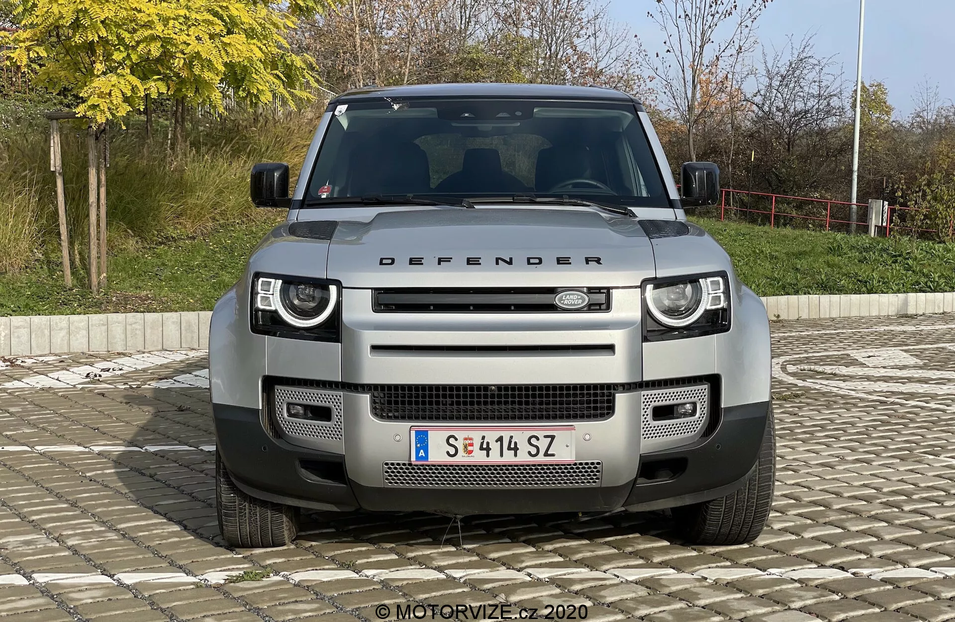 Sprednji pogled na moderni SUV Land Rover Defender 2020, ki prikazuje značilno masko, žaromete in pokrov motorja z napisom 'DEFENDER'. Slika, centralno uokvirjena in posneta pod rahlo dvignjenim kotom, izpostavlja moderne LED žaromete, geometrični vzorec maske, sprednje kamere in senzorje za parkiranje, sprednjo zaščitno ploščo in vlečne točke. Vozilo je postavljeno proti preprostem zunanjem ozadju z grmičevjem in ograjo, pod naravno dnevno svetlobo.