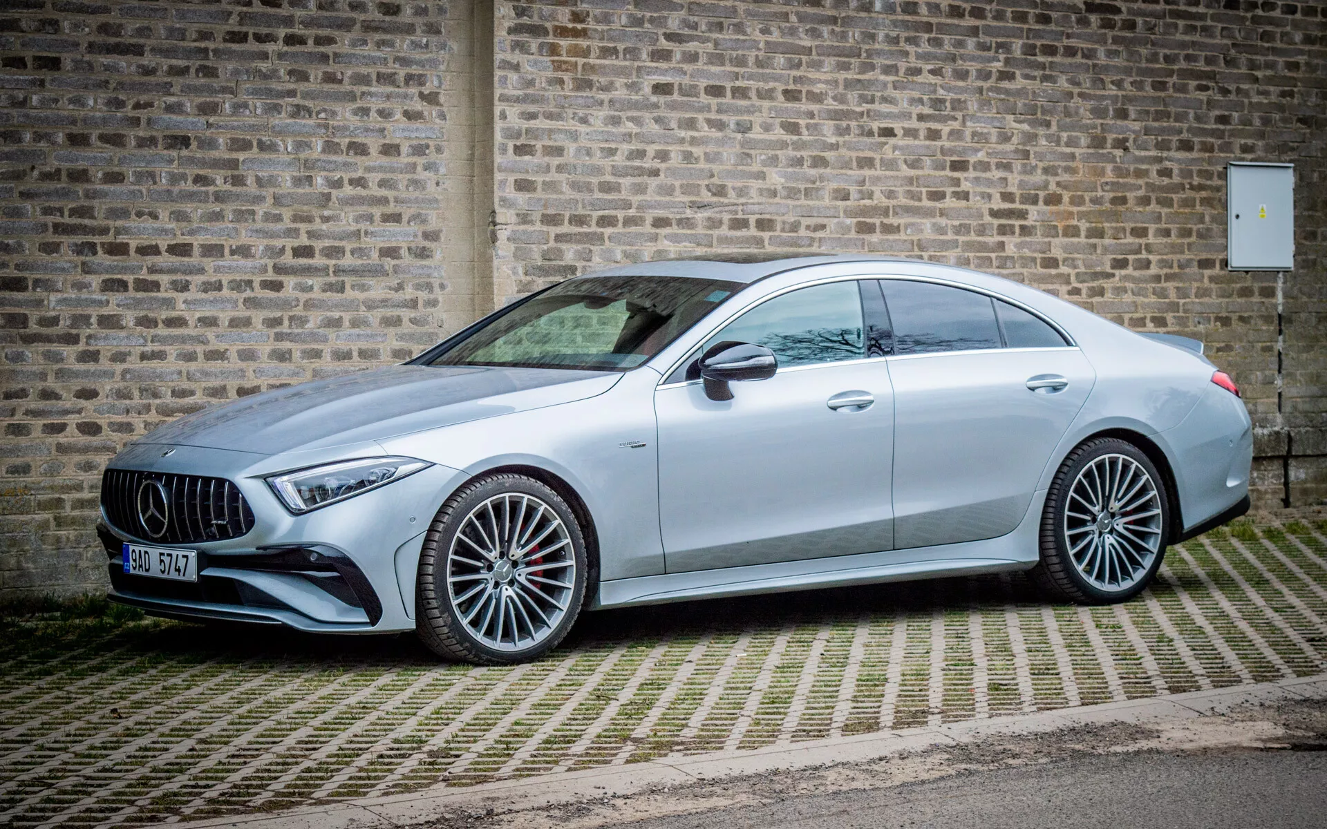 Mercedes-Benz CLS AMG Coupe 2021 iz diagonalne sprednje stranske perspektive, ki prikazuje metalik srebrno barvo, športna večkraka lita kolesa, izstopajočo sprednjo masko z emblemo, LED žaromete in eleganten aerodinamični profil, parkiran na vzorčasti opečni površini ob kamnitem zidu