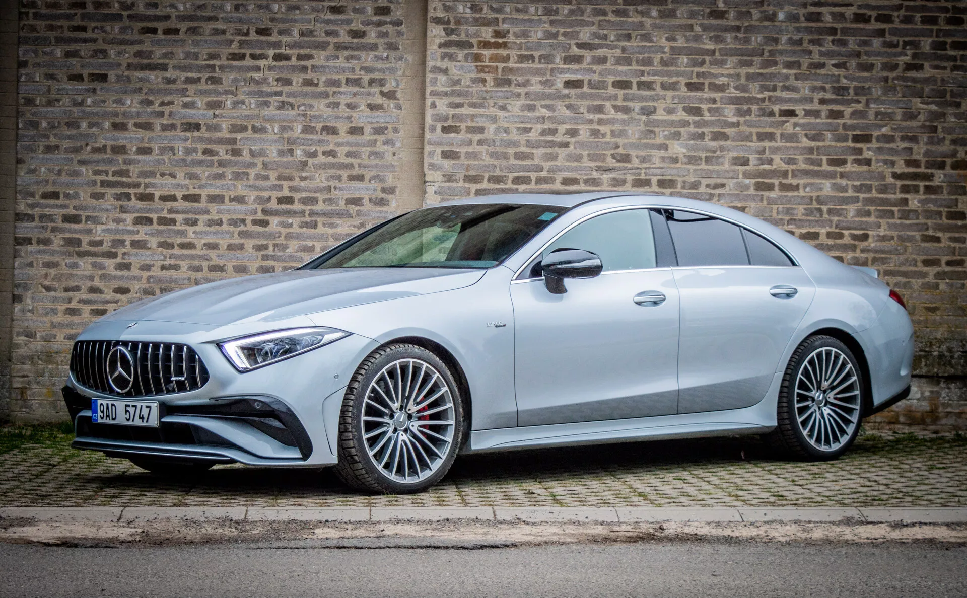 Kupe Mercedes-Benz CLS AMG 2021 v bočnem profilu proti urbanemu ozadju opečnatega zidu, prikazuje LED žaromete, aerodinamično zasnovo in športno silhueto kupeja