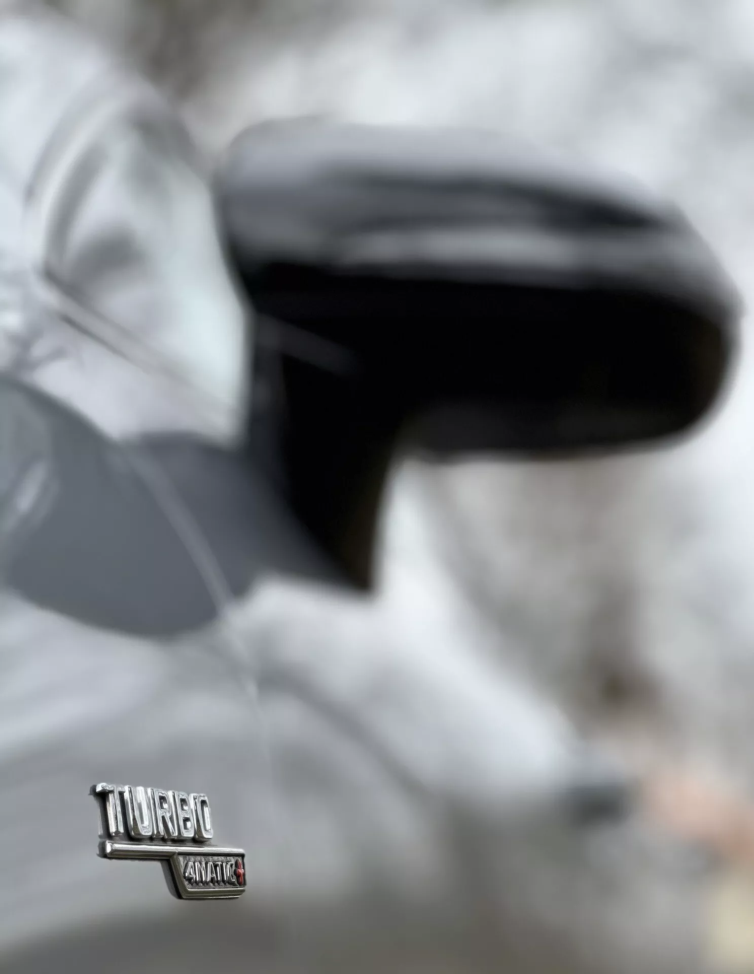 Posnetek emblema Volkswagen Touareg na strani voznika s fokusiranim pogledom na sijoče, aerodinamično stransko ogledalo, kar kaže na sodobno zasnovo vozila in morda napredno tehnologijo, z zamegljenim ozadjem, ki nakazuje, da je avto parkiran.
