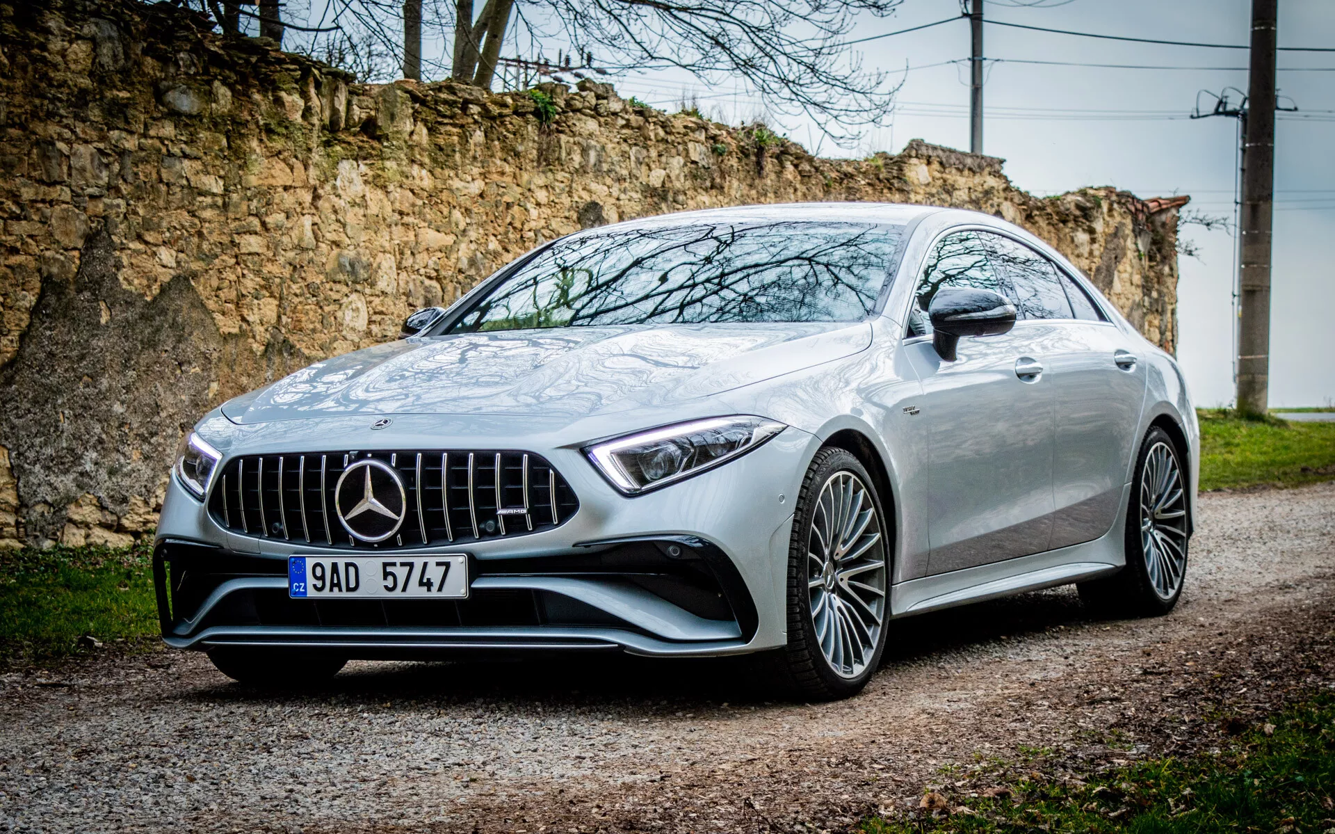 Srebrni kupe Mercedes-Benz CLS AMG 2021 iz trehčetrtinskega sprednjega kota, ki prikazuje rešetko, LED žaromete, sprednji odbijač in platišča iz lahke zlitine na gramozni poti z rustikalnim ozadjem