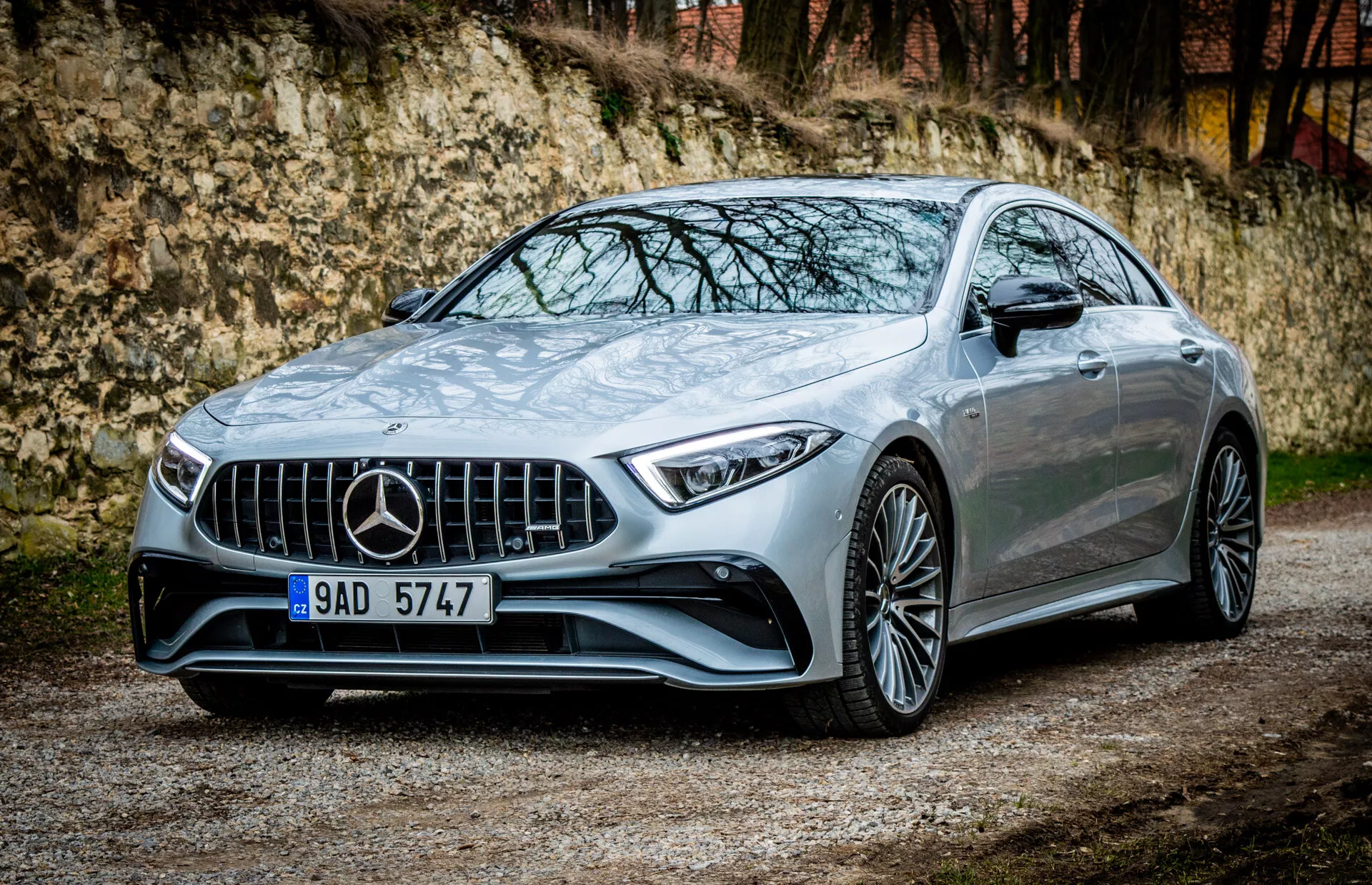 Mercedes-Benz CLS-AMG Coupe leta 2021, parkiran na gramozu z vegetacijo in arhitekturno strukturo v ozadju, prikazuje svojo značilno rešetko, LED žaromete, aerodinamični odbijač, športne linije pokrova, večkraka lita kolesa, vratne kljuke na poteg in potencialno panoramsko streho v luksuzni kovinski obdelavi