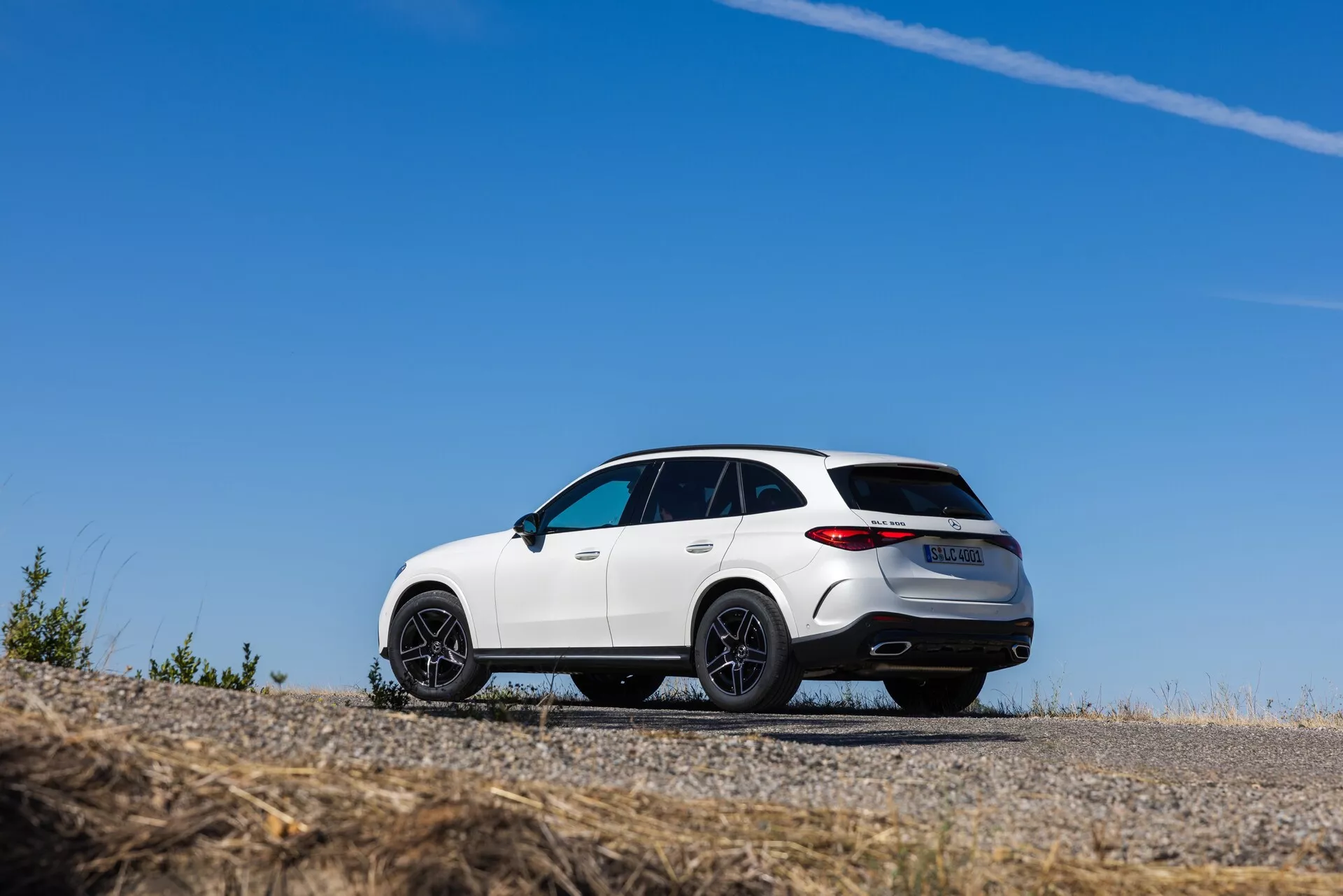 Nizek kot, delni zadnji pogled na bel SUV Mercedes GLC 2022, parkiran na asfaltni površini, ki prikazuje svoj športni dizajn s poševno strešno linijo, velikimi črnimi večkrakimi platišči in LED zadnjimi lučmi. Vozilo ima kromirane dodatke, dvojne izpušne cevi, zadnji spojler, panoramsko streho in zadnji difuzor, poudarja pa njegovo vrhunsko konstrukcijo in zmogljivostni videz.