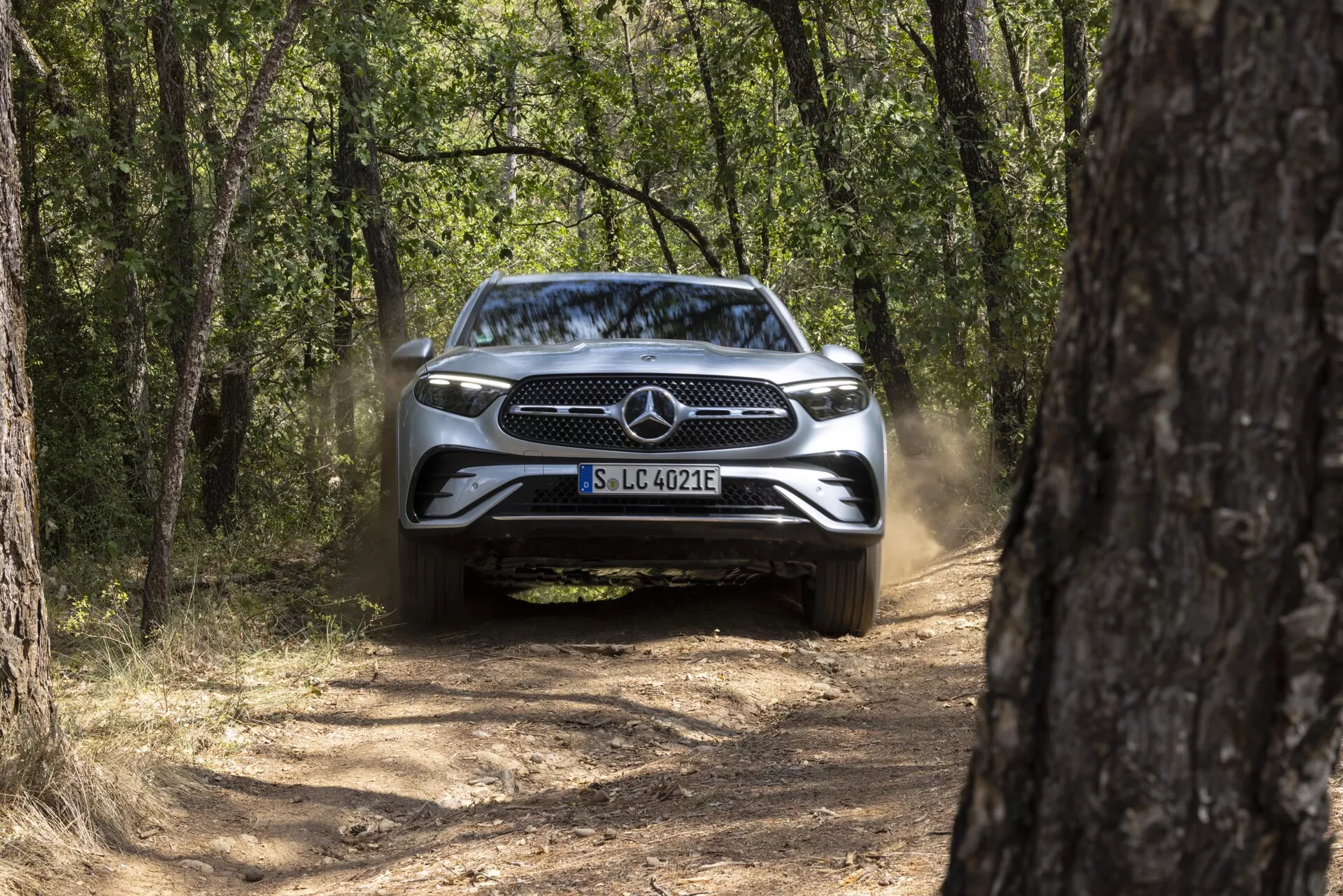 Sprednji pogled na Mercedes GLC 2022 (254) na ozki blatni poti, obkrožen z drevesi, brez vključenih žarometov, prah za njim, sončen dan z naravno svetlobo.