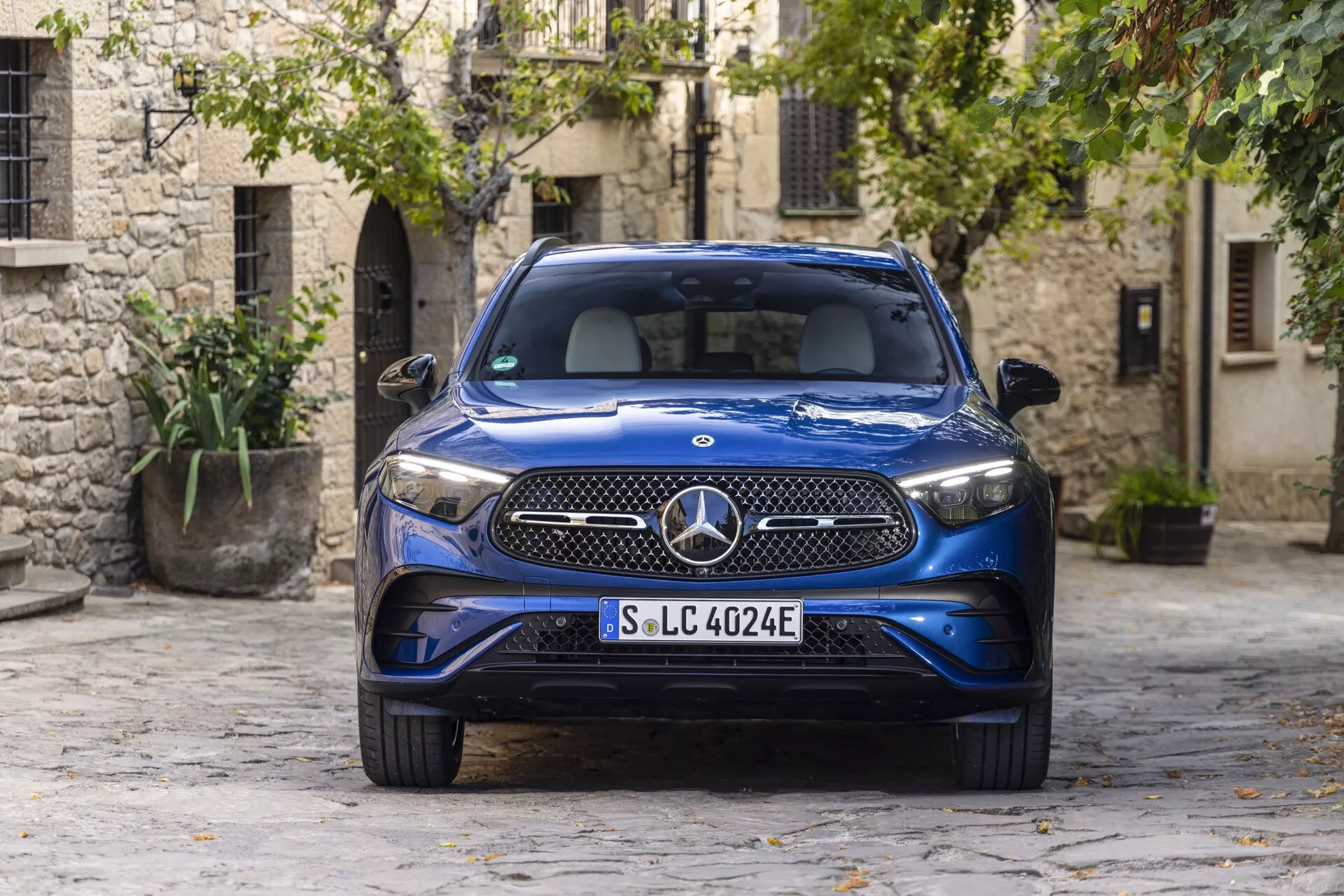 2022 Modri luksuzni SUV Mercedes GLC 254 parkiran v slikovitem, tlakovanem dvorišču starega mesta, ki prikazuje LED žaromete, prepoznavno sprednjo masko z velikim emblemom Mercedes-Benz, značko EQ, ki označuje električno ali priključno hibridno naravo, elegantno kovinsko zunanjost in integrirane zračne odprtine v prednjem odbijaču. Posneto iz sprednjega kota, poudarjajoč sodoben avtomobilski dizajn v primerjavi s tradicionalnimi kamnitimi zgradbami in zelenjem.