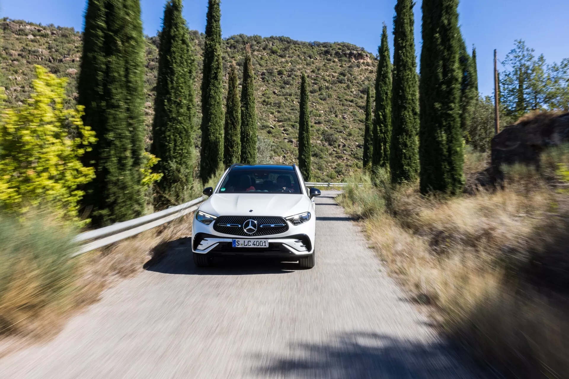 Dinamična slika avtomobila iz sprednje perspektive, Mercedes GLC 2022 (254), ki vozi proti kameri po ozki sončni cesti obdani z visokimi drevesi, prikazuje celotno prednjo fasado, vključno z mrežico z emblemom Mercedes-Benz, elegantnimi LED žarometi, pokrovom motorja, vetrobranskim steklom in delom strehe. Rahla zamegljenost koles in črtasta ozadja, ustvarjena s tehniko zameglitve gibanja, poudarjata premikanje avtomobila in njegove napredne funkcije, kot je možno LED tehnologija žarometov, posodobljena zasnova mrežice z aktivnimi aerodinamičnimi elementi ali senzorji za sisteme za pomoč vozniku in verjetno tudi parkirni senzorji ali kamere.