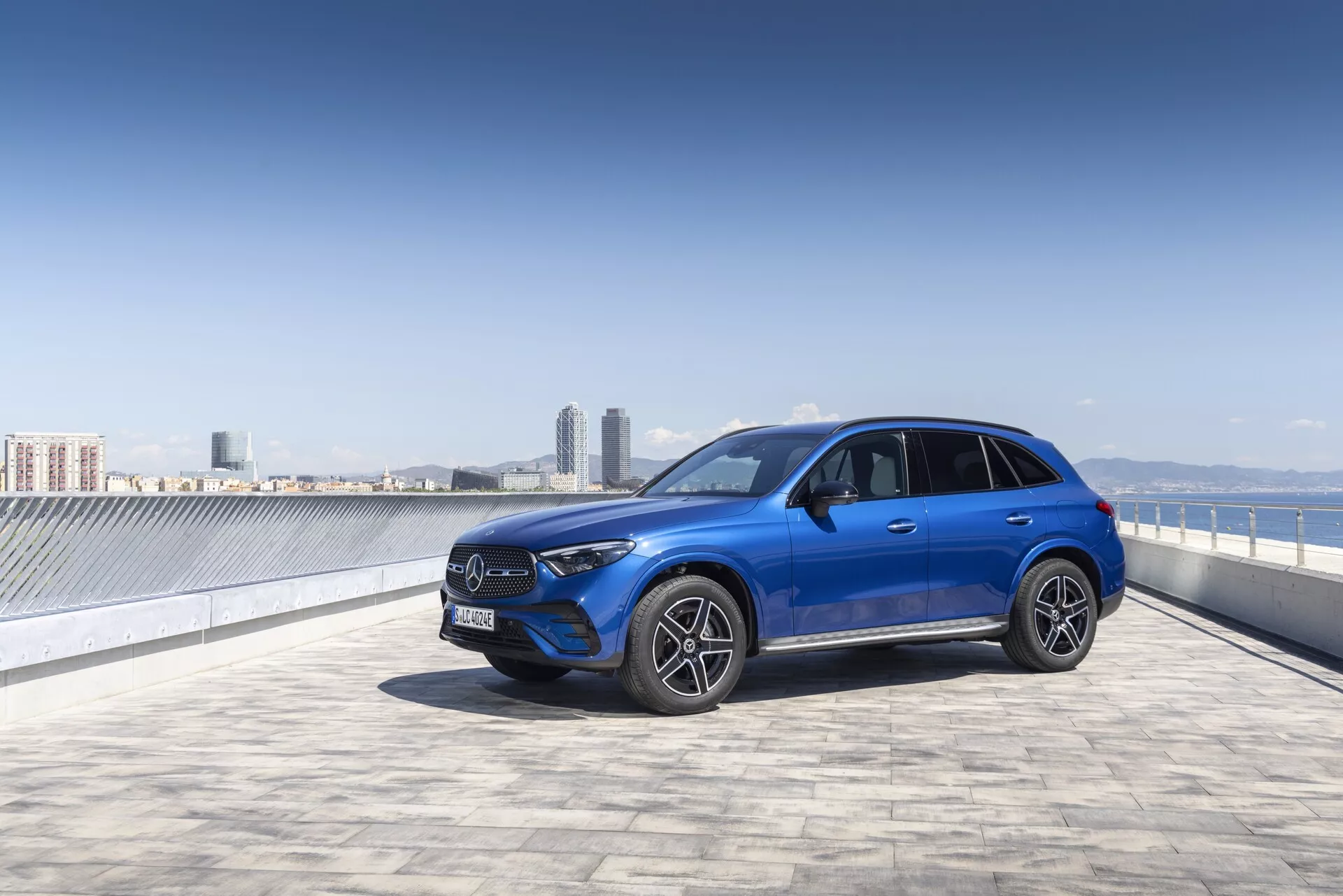 Svetlo modri SUV Mercedes GLC 2022 na strešnem parkirišču s čistim nebom in mestno silhueto v ozadju, zajet iz treh četrtin sprednje strani. Sliko poudarja izrazita sprednja maska z velikim emblemom, kotne LED žaromete, sprednji odbijač z možnimi senzorji, večkraka aluminijasta platišča in stranska ogledala z vgrajenimi smerniki. Elegatne linije in športna drža avtomobila so dobro poudarjene, kar nakazuje na napredno tehnologijo in urbano prefinjenost.