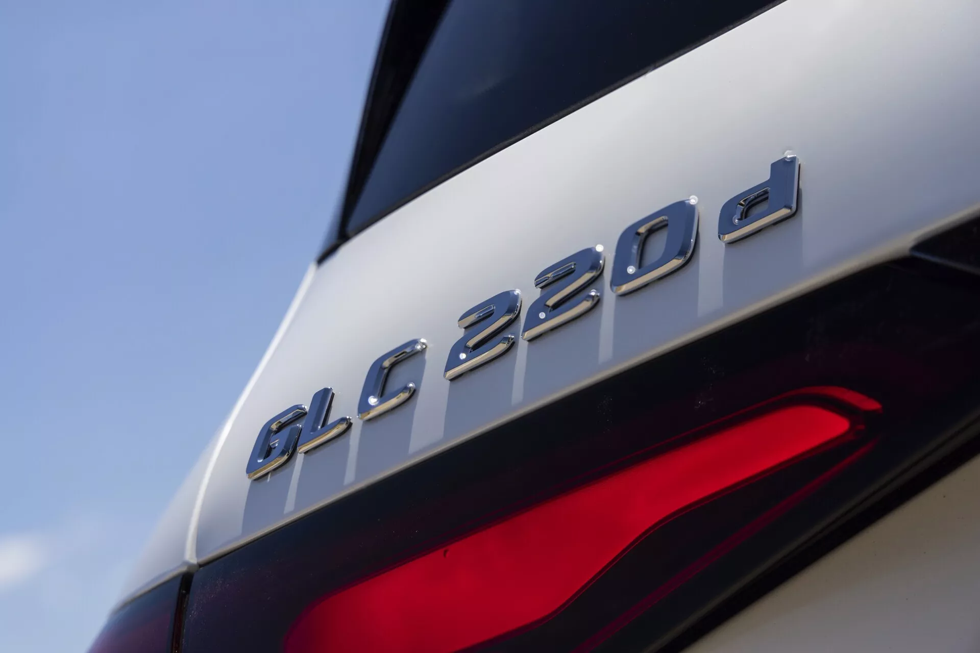 Povečana slika zadnjega dela Mercedes-Benz GLC 220d 2022, ki poudarja značko modela tik nad delno vidno zadnjo lučjo. Slika se osredotoča na značko 'GLC 220d', kar pomeni, da gre za dizelsko različico. Posnetek iz nizkega, rahlo navzgor usmerjenega kota prikazuje elegantne oblikovalske linije, kovinsko obdelavo značke v kontrastu s sijočim lakom ter sodobne LED elemente zadnje luči, kar kaže na napredne svetlobne tehnologije.