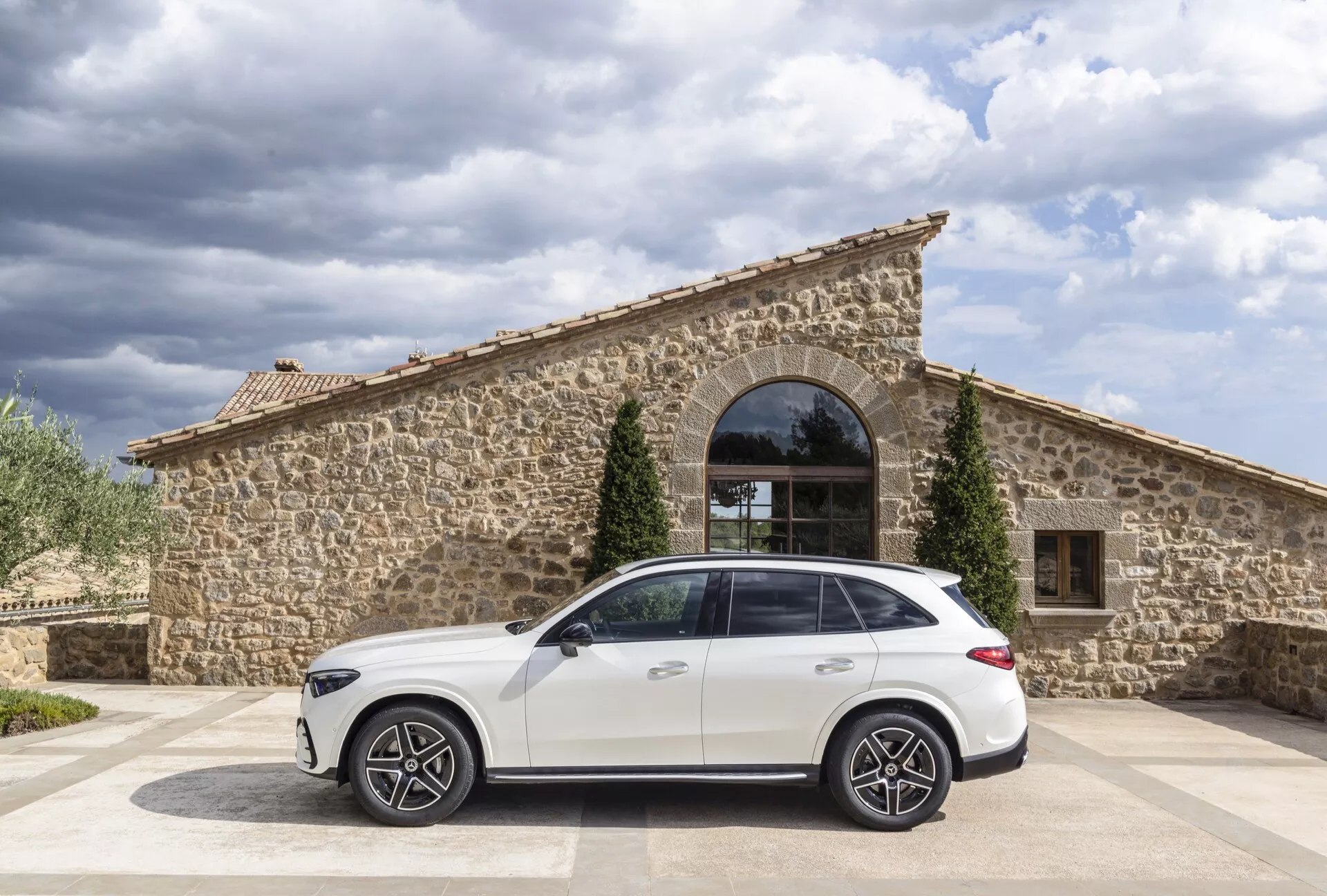 Stranski profil belega SUV Mercedes GLC 2022, parkiranega na tlakovani površini z rustikalno kamnito zgradbo z velikim obokanom oknom v ozadju. Oblačno nebo omogoča naravno dnevno svetlobo, kot kamere prikazuje streho in linije karoserije avtomobila z desne strani. Opazne lastnosti vključujejo velika aluminijasta platišča z več kraki, značilno linijo pod okni, vgrajene kljuke vrat, strešne nosilce in anteno v obliki plavuti morskega psa.