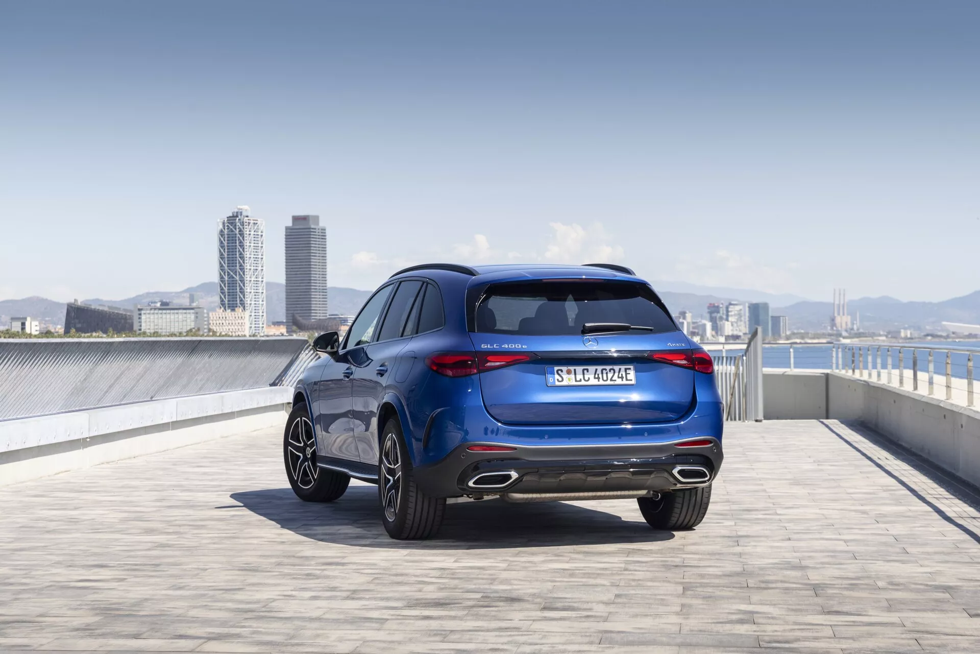 Nizek kot posnetka Mercedesa GLC 2022 (254) iz zadnjega tretjinskega kota, ki prikazuje njegove elegantne in športne oblikovne linije, zadnje luči, aluminijasta platišča, kovinsko barvo in znamko. Slika zajema celotno višino in širino vozila, izpostavlja pa elemente, kot so zadnje steklo, izpušni sistem, registrska tablica in morebitna oznaka nivoja opreme ali pogona. Zunanje okolje, verjetno strešno parkirišče z urbano silhueto v ozadju, poudarja sodoben in prefinjen videz vozila.