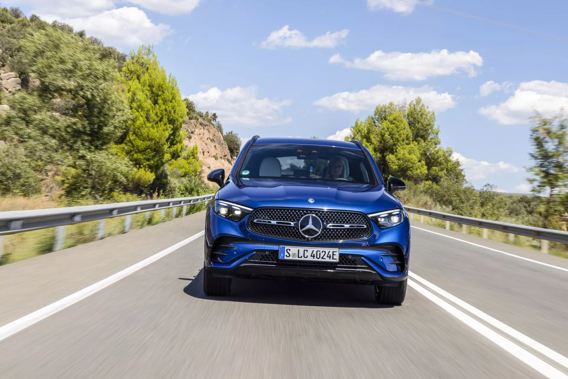 Slika modrega modela Mercedes-Benz GLC 254 iz leta 2022, posneta iz sprednjega tričetrtinskega kota, poudarja eleganten dizajn, izrazit trikraki zvezdni emblem, podroben vzorec v rešetki, elegantne žaromete, ki se zlijejo z blatniki in jasne podrobnosti sprednjega odbijača, pokrova motorja in vrat. Avto, fotografiran v gibanju na slikoviti dvosmerni cesti z zelenimi hribi v ozadju, verjetno vključuje električno ali hibridno tehnologijo, kar nakazuje oznaka 'EQ' na nosilcu registrske tablice. Slika je podrobna z zamegljenostjo gibanja, ki kaže na gibanje, in prikazuje dnevne luči avtomobila podnevi pod jasnim nebom.