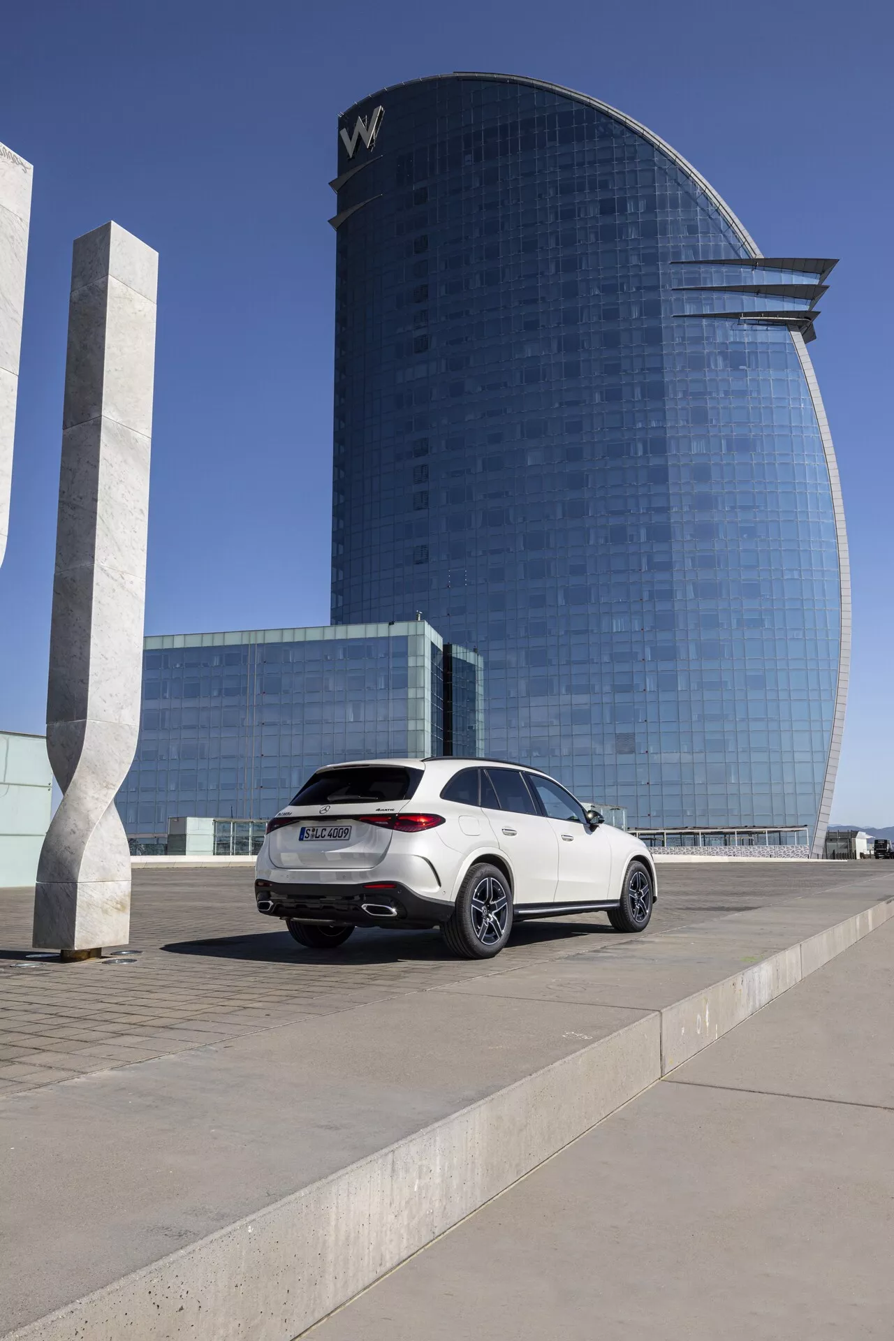 Mercedes GLC 2022 - Beli luksuzni SUV parkiran na trgu; srednje-nizek kot, ki prikazuje stranski profil na strani potnika in zadnji del, prikazuje sodobne zadnje luči, dvojni izpušni sistem, večkraka aluminijasta platišča, strešno linijo podobno kupeju, zatemnjena okna; v ozadju sodobna hotelska ali poslovna stavba z odsevno stekleno fasado v jasni dnevni svetlobi.