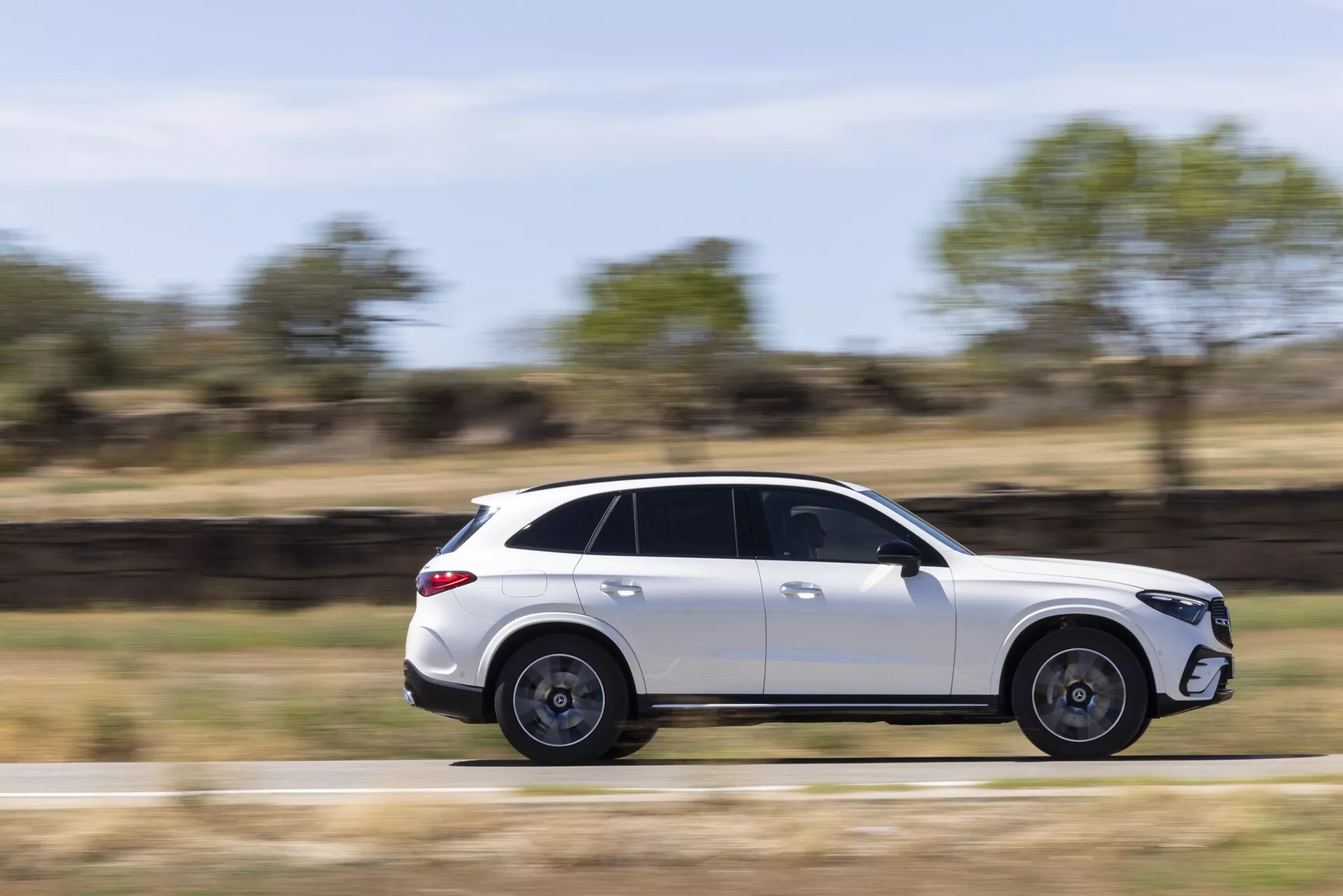 Slika modernega, elegantnega belega SUV-ja Mercedes GLC 2022 v gibanju, posneta s stranskega profila. Vozilo je zajeto s tehniko panninga, ki ga ohranja ostrega, medtem ko ozadje ni ostro, kar prikazuje gibanje. S toniranimi okni, strešnim oknom, petkrakovimi aluminijastimi platišči in aerodinamično zasnovo je ta SUV postavljen v podeželsko okolje z naravno dnevno svetlobo.