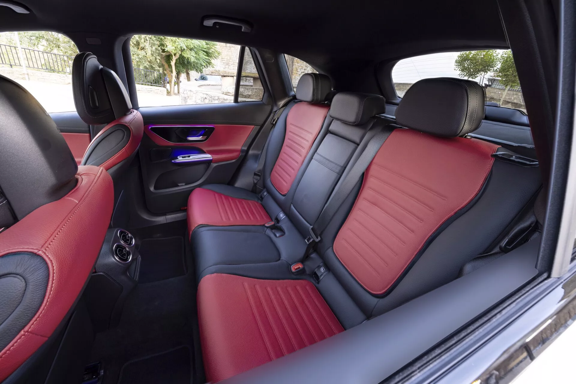 Pogled na notranjost Mercedes GLC 2022 z desne strani, ki zajema zadnje sedeže z luksuznim dvobarvnim oblazinjenjem v rdeči in črni barvi, iz usnja ali imitacije usnja z natančnim šivom. Funkcije vključujejo zložljivo sredinsko naslonjalo za roke s držali za pijačo, modro LED osvetlitev, ki izboljšuje sodobno atmosfero kabine, zadnjo klimatsko odprtino ter integrirane zvočnike v vratnih ploščah, kar kaže na napredno osvetlitev, luksuzno obdelavo in udobje za potnike.