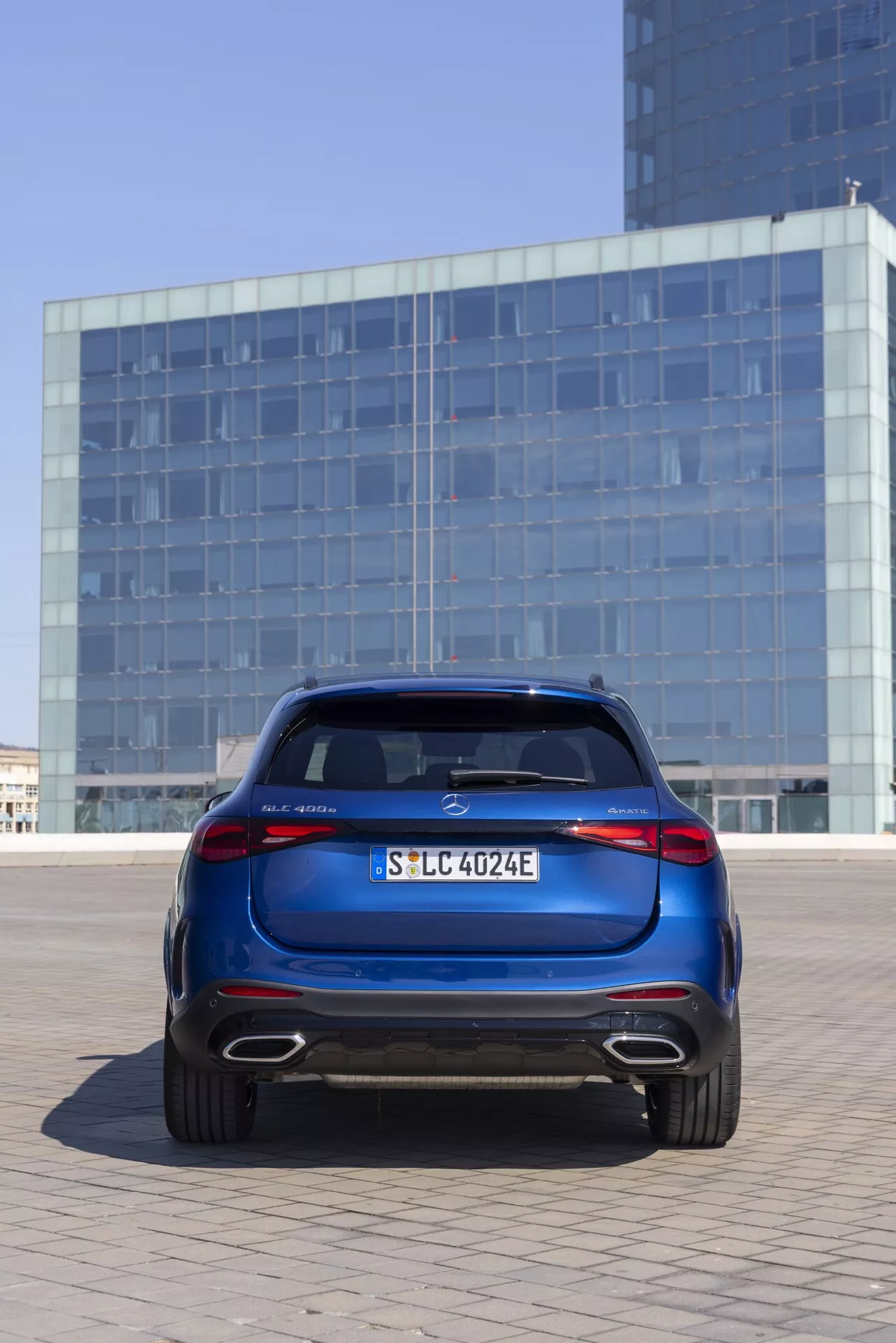 Zadnji pogled na Mercedes GLC 2022 (254) v globoki kovinski modri barvi, postavljen na prostem v mestnem okolju z odsevno stekleno fasado stavbe v ozadju. Avto je centriran iz rahlo nizkega kota, kar poudarja prtljažna vrata, zadnji odbijač, LED zadnje luči, aluminijasta platišča in strešni spojler. Funkcije vključujejo izpostavljen sredinski značko, registrsko tablico, oznako modela na levi, 'E' kar označuje električni ali hibridni model na desni, in zadnji difuzor z izpušnimi odprtinami, kar nakazuje hibridni model.