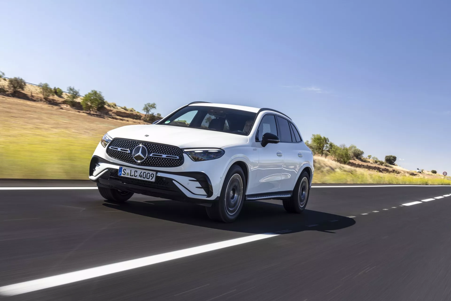 Dinamičen posnetek luksuznega SUV-ja Mercedes GLC 2022, ki se premika naprej po gladki asfaltirani cesti, prikazuje njegov sprednji in stranski profil. Značilnosti vključujejo ikonično masko z emblemom z zvezdo s tremi kraki, elegantne LED žaromete, senzorje na sprednjem odbijaču, ki nakazujejo napredne sisteme za pomoč vozniku, oblikovane linije pokrova motorja, aluminijasta platišča, aerodinamična stranska ogledala z integriranimi utripalkami in tekočo linijo strehe za bolj športno silhueto. Dominantna drža vozila in rahla zamegljenost okoli koles poudarjajo gibanje. Ozadje je jasno nebo in puščavska pokrajina.