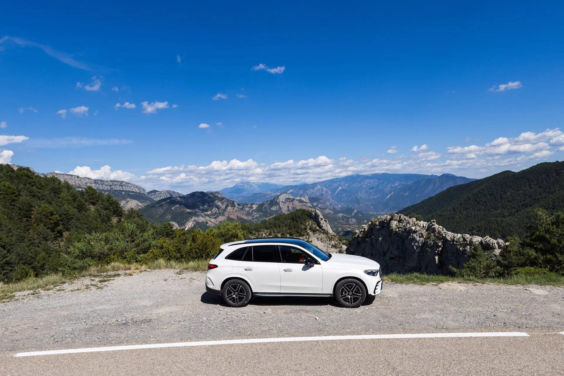 Bel, športni SUV Mercedes GLC 2022 je parkiran na ravni, asfaltirani površini, obdani z naravno gorsko pokrajino, pod jasnim modrim nebom. Bočni profil prikazuje voznikovo stran vozila in izpostavlja oblikovalske elemente, kot so aluminijasta kolesa, stranska ogledala, obrobe oken in linije karoserije. Osvetlitev in jasnost omogočata vidnost subtilnih kontur na stranskih vratih.