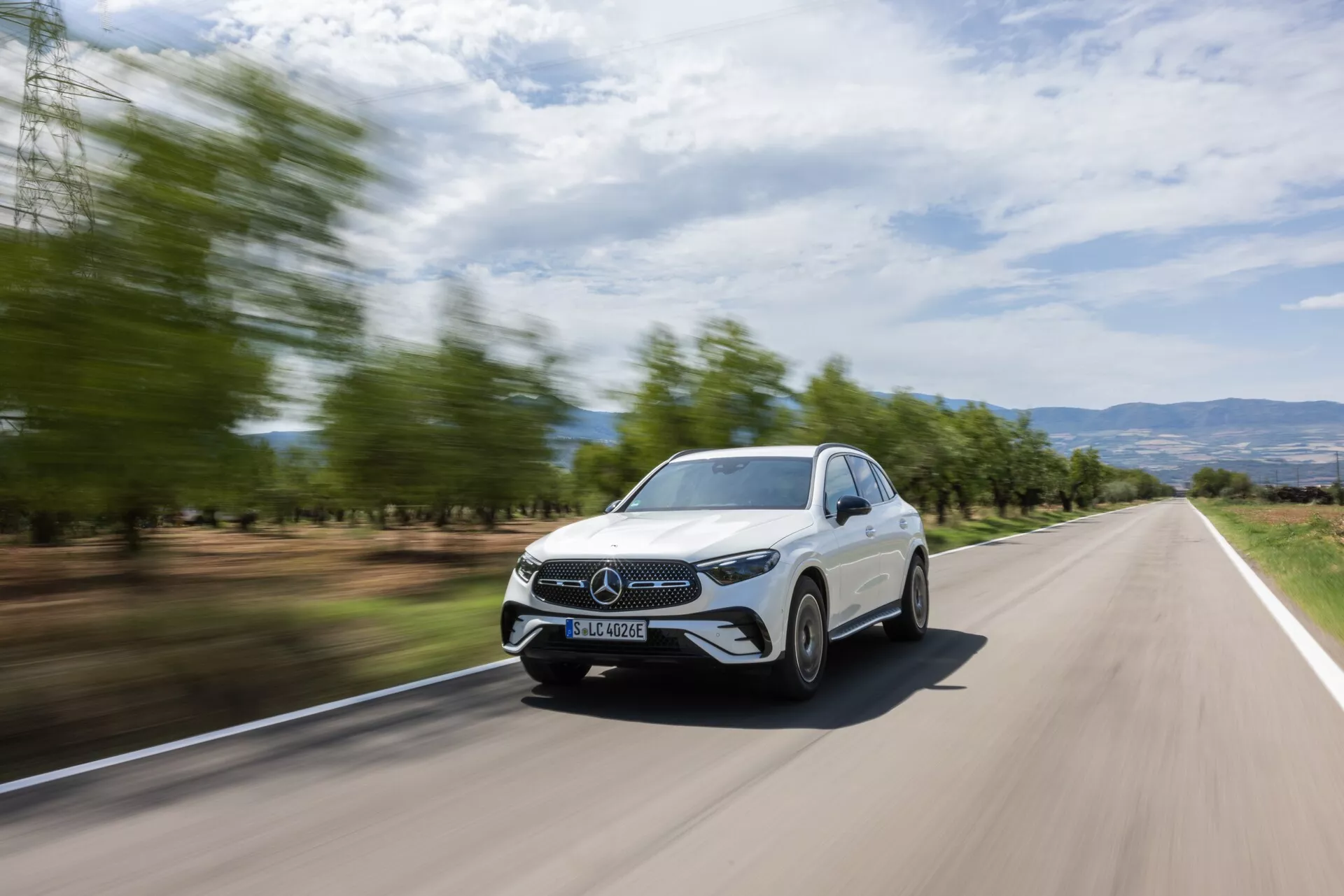 Sprednji trikotni pogled na bel Mercedes GLC 2022 v gibanju na dvopasovni cesti, prikazuje njegovo sprednjo masko, žaromete, del pokrova motorja, vetrobransko steklo in voznikovo stran. Ozadje je zaradi gibanja zamegljeno, kar poudarja hitrost avtomobila. Eleganten dizajn, zaprta maska, energetsko učinkoviti LED žarometi in vidne dnevne luči, skupaj s sodobno pokrajino z zamegljenimi drevesi in gričevnatim terenom, predstavljajo dinamičen in stilski videz, idealen za promocijsko gradivo.