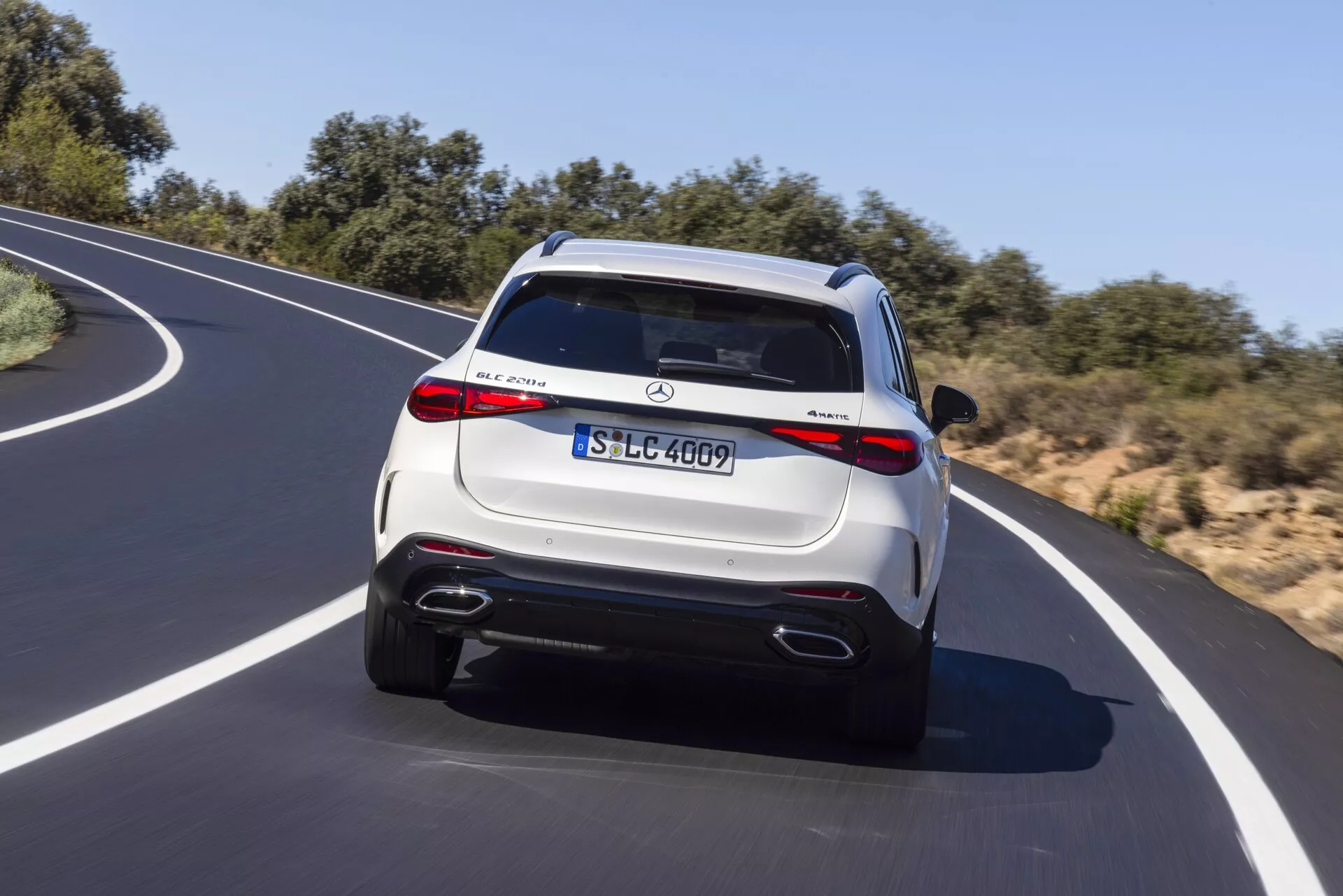 Pogled na zadnji del luksuznega SUV vozila Mercedes-Benz GLC 2022 v gibanju po zaviti asfaltni cesti pod jasnim nebom. Vozilo ima kovinsko barvo svetlega odtenka, LED luči, dvojni izpušni cevi, zadnji odbijač s difuzorjem in strešni spojler z integrirano zavorno lučjo. Vidni so prominentni logotipi in evropska registrska tablica, ki označujejo model in podrobnosti opreme. Večkraki platišči in možne visokotehnološke funkcije, kot so parkirni senzorji in vzvratna kamera, dodajajo luksuzni pridih.