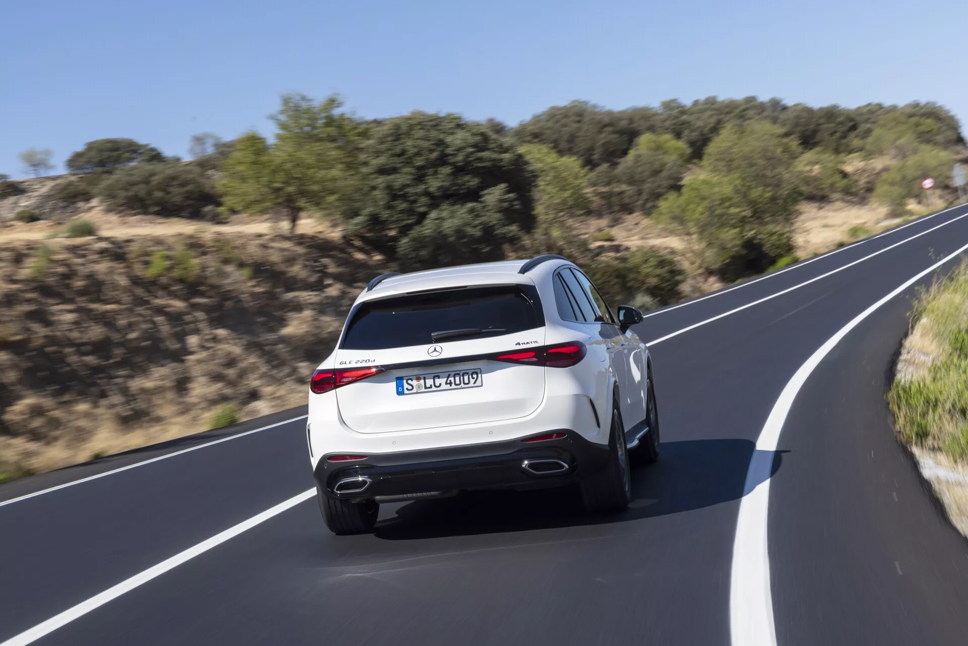 Zadnji pogled na sodoben Mercedes GLC SUV 2022 v gibanju, ki prikazuje dvojne izpušne cevi, aerodinamični zadnji difuzor, LED zadnje luči, športni zadnji spojler in praktične funkcije, kot so zadnji brisalec in visoko nameščena tretja zavorna luč. Svetlo barvana barva SUV-ja, kot je bela ali srebrna, poudarja njegov vrhunski videz med vožnjo po gladki asfaltirani cesti, obkroženi z naravno vegetacijo. Zameglitev gibanja poudarja dinamične zmogljivosti vozila.