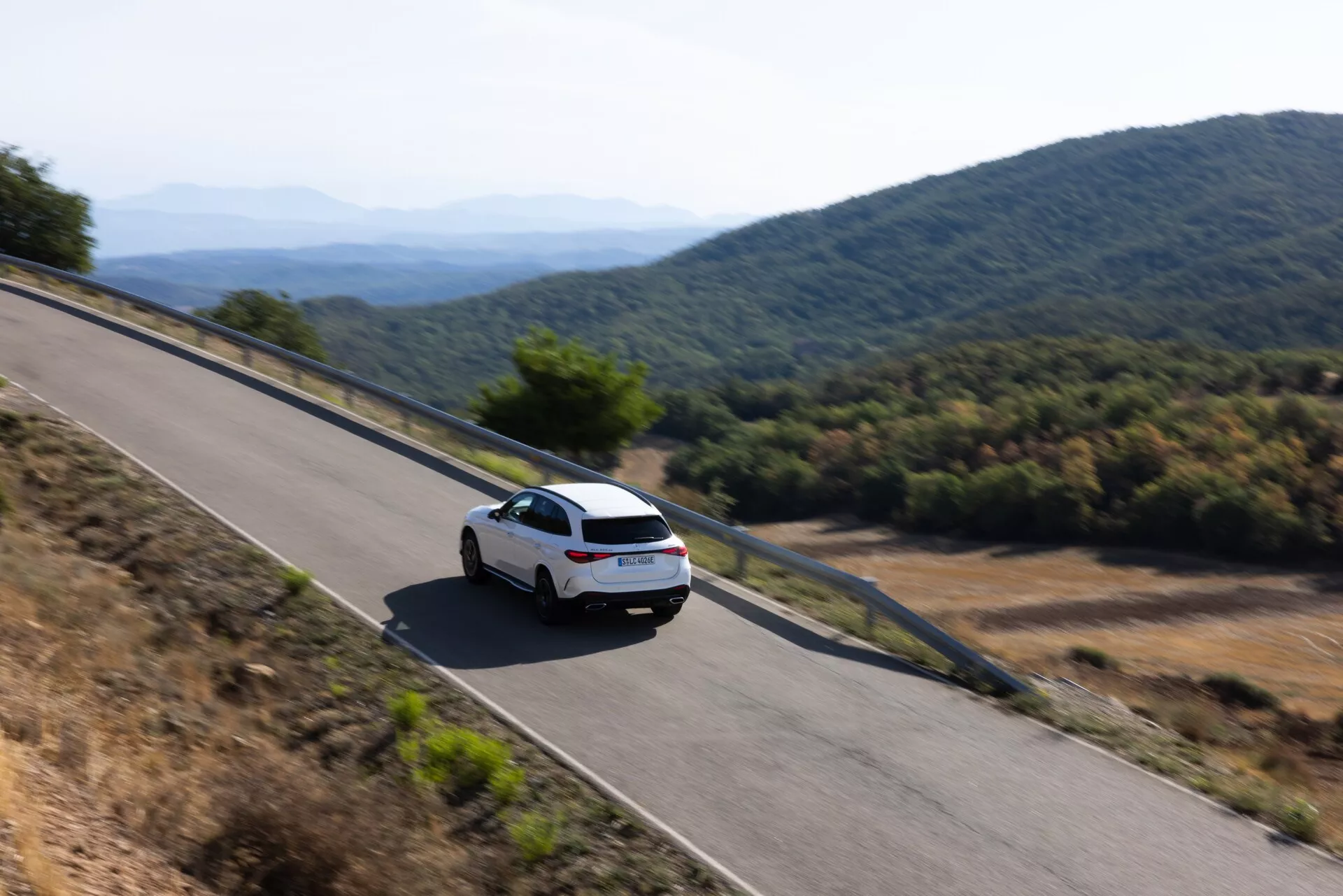 Vrhunski zadnji trikotni pogled na sodobni beli Mercedes GLC 2022 (254) vozeči po gorati cesti. Avto, prikazan z jasnim zadnjim vetrobranskim steklom, zadnjimi lučmi in delom prtljažnika, zajema občutek gibanja z rahlim zamegljevanjem, obkrožen z naravno in toplo svetlobo bodisi zjutraj ali pozno popoldne.