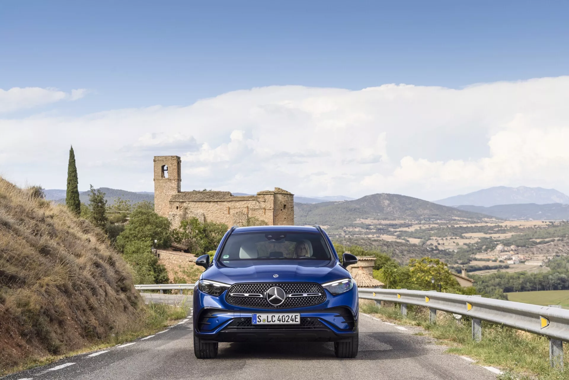 Mercedes-Benz GLC 254 iz leta 2022 viden pod kotom z vidnim sprednjim in stranskim delom. Avto ima elegantne LED žaromete, izrazito rešetko in športna platišča iz zlitine. Parkiran na mestni ulici z modernimi stavbami v ozadju, pri svetlih dnevnih razmerah, ki poudarjajo njegovo kovinsko srebrno barvo.