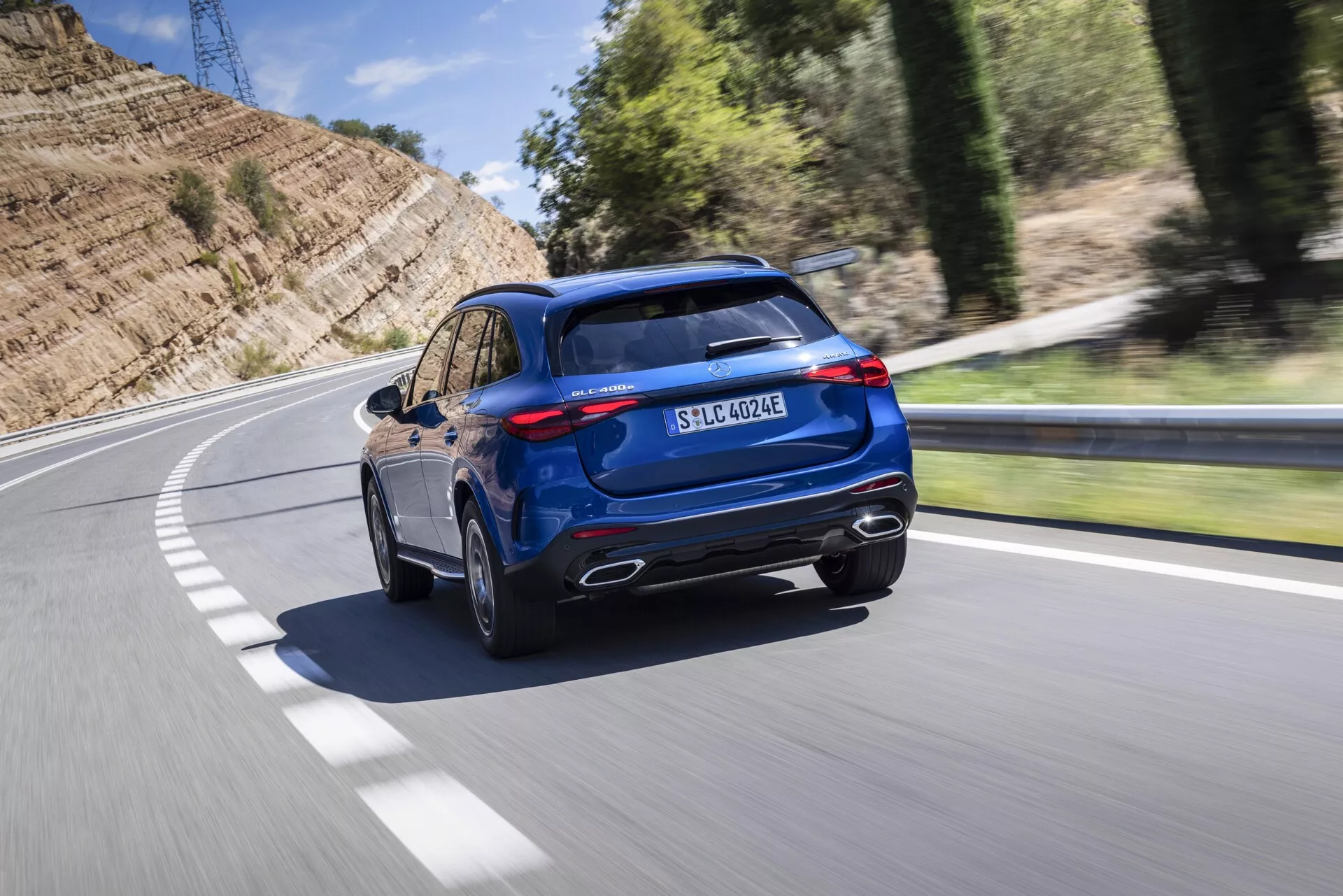 Slika prikazuje dinamičen zadnji tridelni pogled modrega terenskega vozila Mercedes GLC 2022, ki vozi po asfaltirani cesti, ki se zavija v levo. Zajemano iz rahlo povišanega kota, značilnosti vozila vključujejo kromirane izpušne cevi, robusten zadnji odbijač s črnimi oblogami, elegantne LED zadnje luči in strmo nagnjeno zadnje steklo. Športni zadnji spojler, panoramska streha in vidne strešne nosilce povečujejo prestižni videz. Bočni profil razkriva večkrake litinske platišča in pnevmatike z nizkim profilom, kar poudarja cestne zmogljivosti. Polnilna postaja pred zadnjo lučjo in značko EQ ponazarjata možnosti priključnega hibrida ali električnega pogona, medtem ko značko 4MATIC označuje štirikolesni pogon. Zamegljeno ozadje zaradi gibanja nakazuje, da je vozilo v gibanju in daje občutek hitrosti in dinamike.