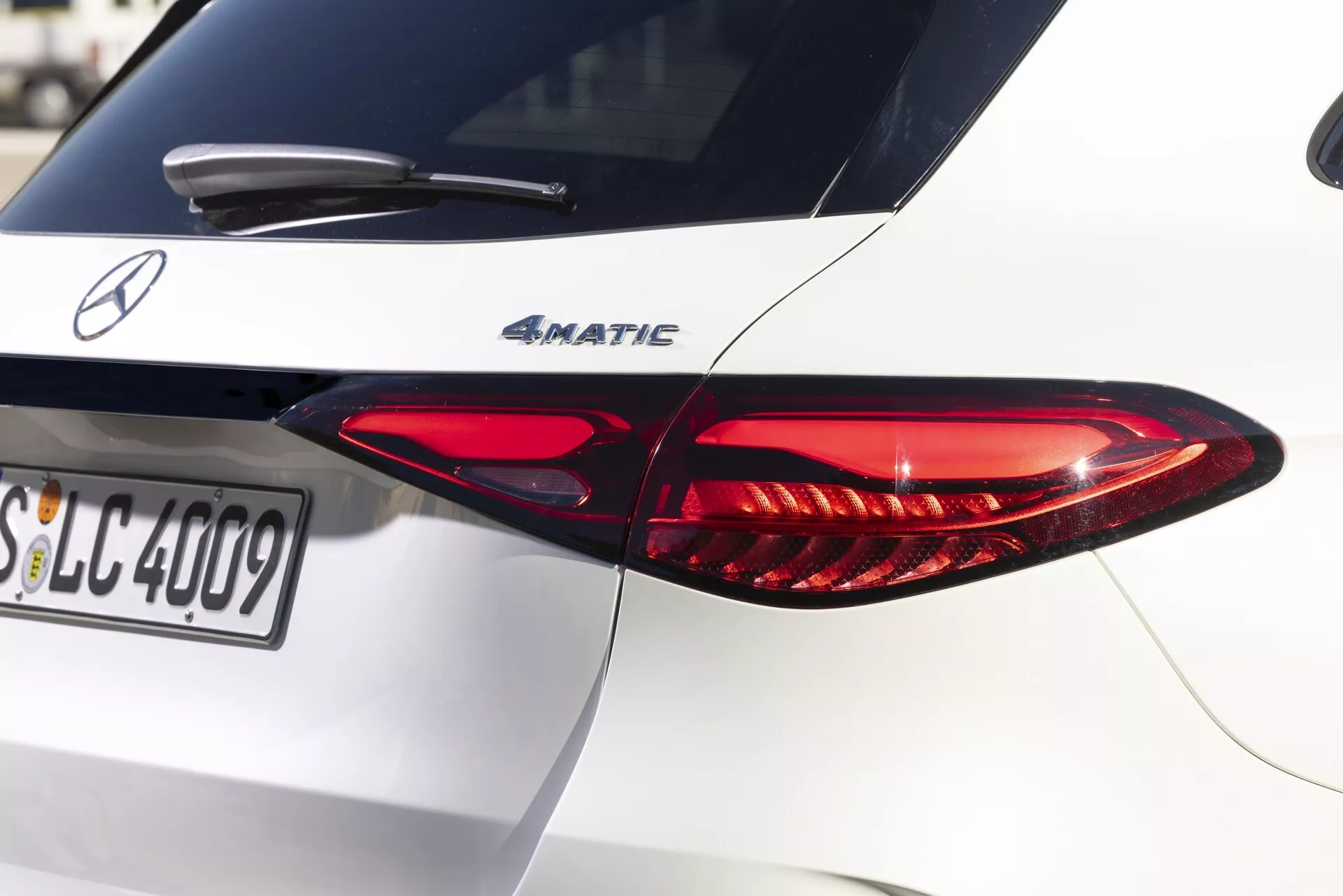 Pogled od blizu zadnjega desnega dela belega Mercedes-Benz GLC 2022, ki poudarja značilno podolgovato zasnovo zadnje LED luči. Zadnja luč ima rdeče in prozorne odseke, ki vsebujejo različne svetlobne funkcije. Pokrov prtljažnika prikazuje oznako modela '4MATIC', kar pomeni štirikolesni pogon. Naravna sončna svetloba meče sence na dobro vzdrževano karoserijo avtomobila, ki se osredotoča na zadnjo luč in značko s plitvo globinsko ostrino. Vidna registrska tablica se začne s 'SLC'. Visokokakovostna slika, ki zajema drobne podrobnosti teksture barve in jasnosti besedila, idealna za prikaz avtomobilskega oblikovanja in svetlobne tehnologije.