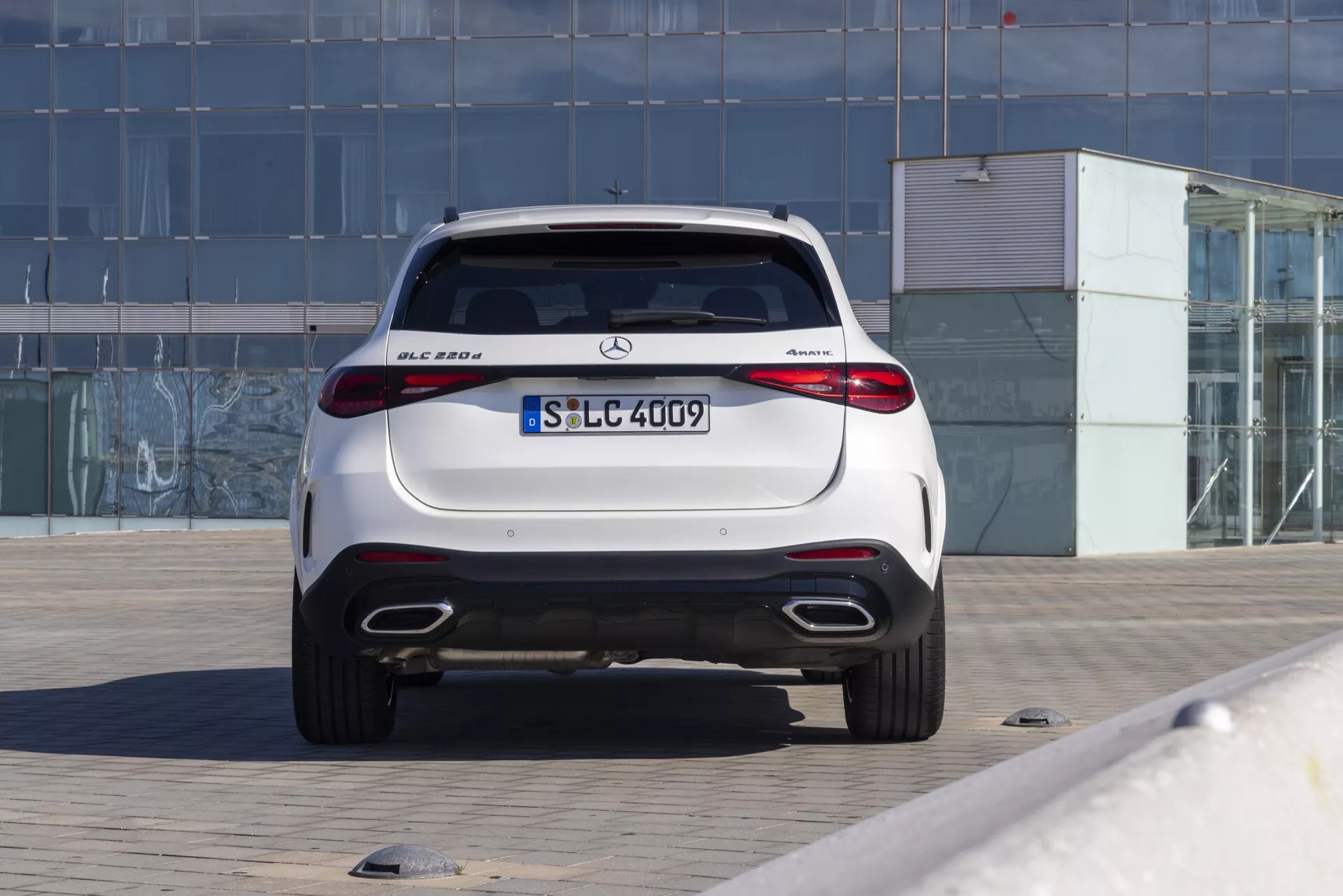 Zadnji pogled na luksuzni kompaktni SUV Mercedes-Benz GLC 300 iz leta 2022 s pogonom na vsa kolesa 4MATIC, parkiran na tlakovani površini. Fotografija prikazuje aerodinamična zadnja vrata prtljažnika, integriran zadnji spojler, LED zadnje luči z vodoravno zasnovo, dvojne izpušne cevi, vgrajene parkirne senzorje, zadnji difuzor in sijajni zaključek. Avto je v središču okvirja z ozadjem, ki vključuje stavbo s stekleno fasado, kar kaže na urbano ali poslovno okolje. Dobro osvetljena slika poudarja elegantne in športne oblikovne značilnosti, značilne za moderna vozila Mercedes-Benz.