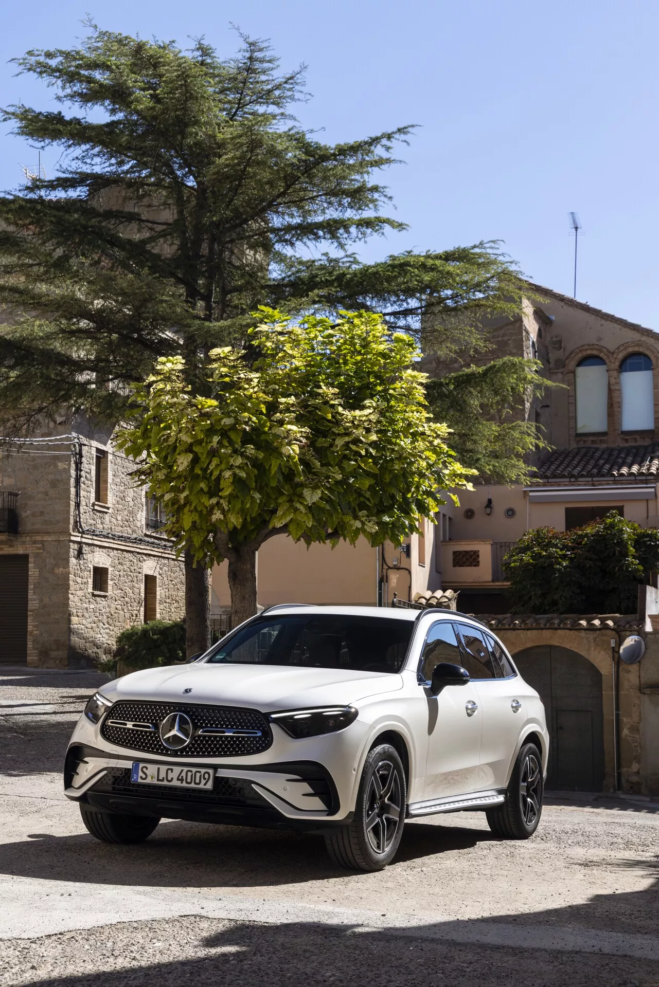 Bel Mercedes-Benz GLC 2022 (254) SUV parkiran na kockasti ulici v zgodovinskem evropskem mestu, zajet iz frontalnega trikotnega kota. Vidne značilnosti vključujejo velik znak Mercedes-Benz na mreži, LED žaromete, aluminijasta platišča, možne kamere/senzorje na sprednjem odbijaču, aerodinamično zasnovo karoserije in ogledala, ki bi lahko imela dodatne kamere ali indikatorje smeri. Okolje in naravna svetloba poudarjata razkošen videz vozila.