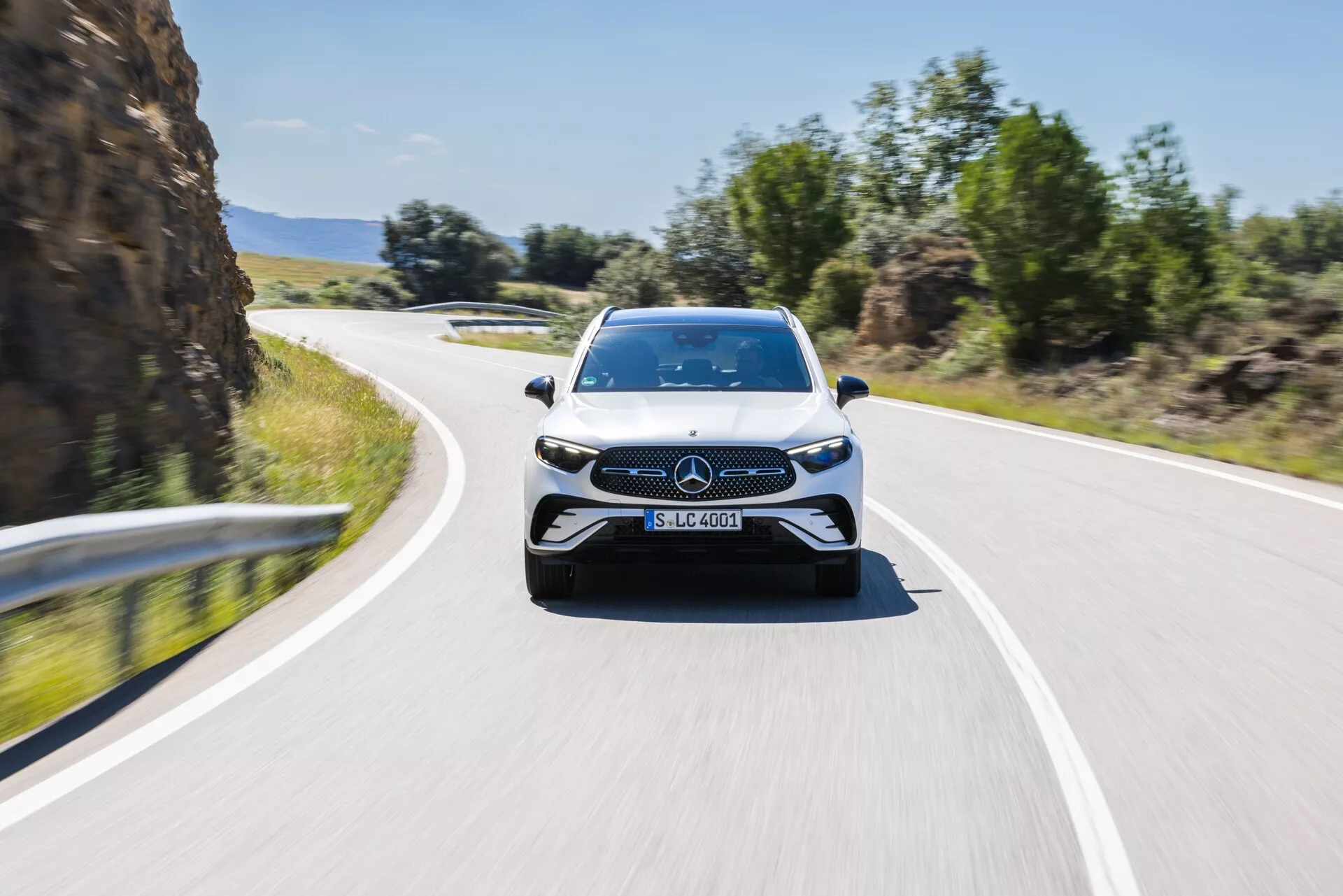 Spredaj pogled na gibajoči se Mercedes-Benz GLC 2022, ki je zajet nekoliko izven centra in prikazuje sprednji del ter del stranic vozila. Slika poudarja emblem in eleganten dizajn rešetke, LED žaromete, velike dovode zraka in oblikovne linije pokrova motorja, kar nakazuje športni in moderni dizajn. Zamegljeno ozadje kaže na občutek hitrosti, saj vozilo vozi po dvosmerni cesti pod čistim nebom, kar poudarja njegove dinamične in napredne tehnološke lastnosti.