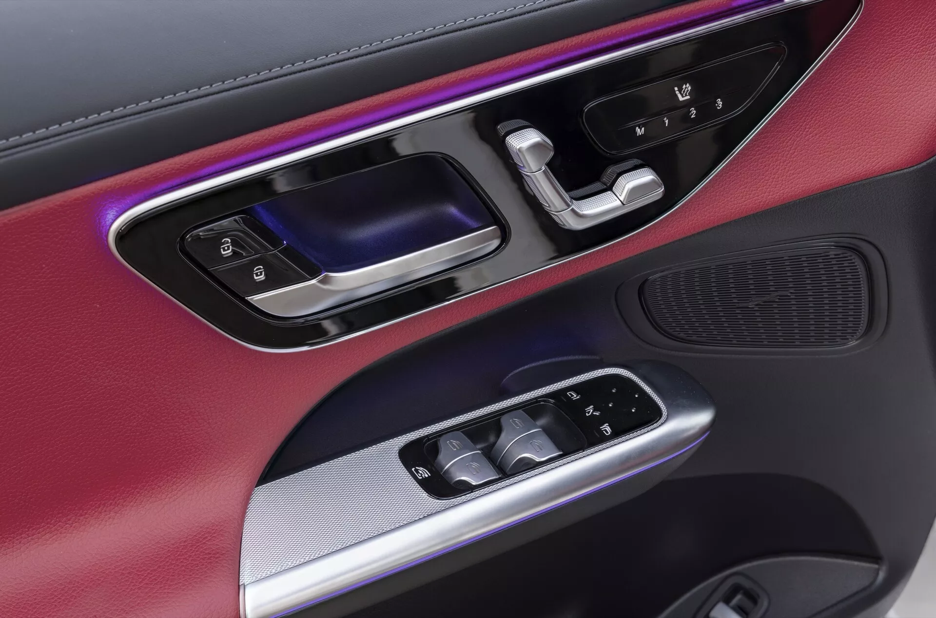 Bližnji posnetek voznikovega vratnega panela Mercedesa GLC (254) iz leta 2022, ki prikazuje mešanico materialov, vključno z kovinskimi oblogami in mat črnimi zaključki. Vključuje barvno ambientalno osvetlitev, ergonomsko oblikovan ročaj v sijajni črni in kovinski barvi, osvetljene električne kontrole oken, električno nastavljiva bočna ogledala in visoko kakovostno zvočno mrežo, vgrajeno na dnu, kar poudarja luksuzno in tehnološko napredno notranjo zasnovo avtomobila.