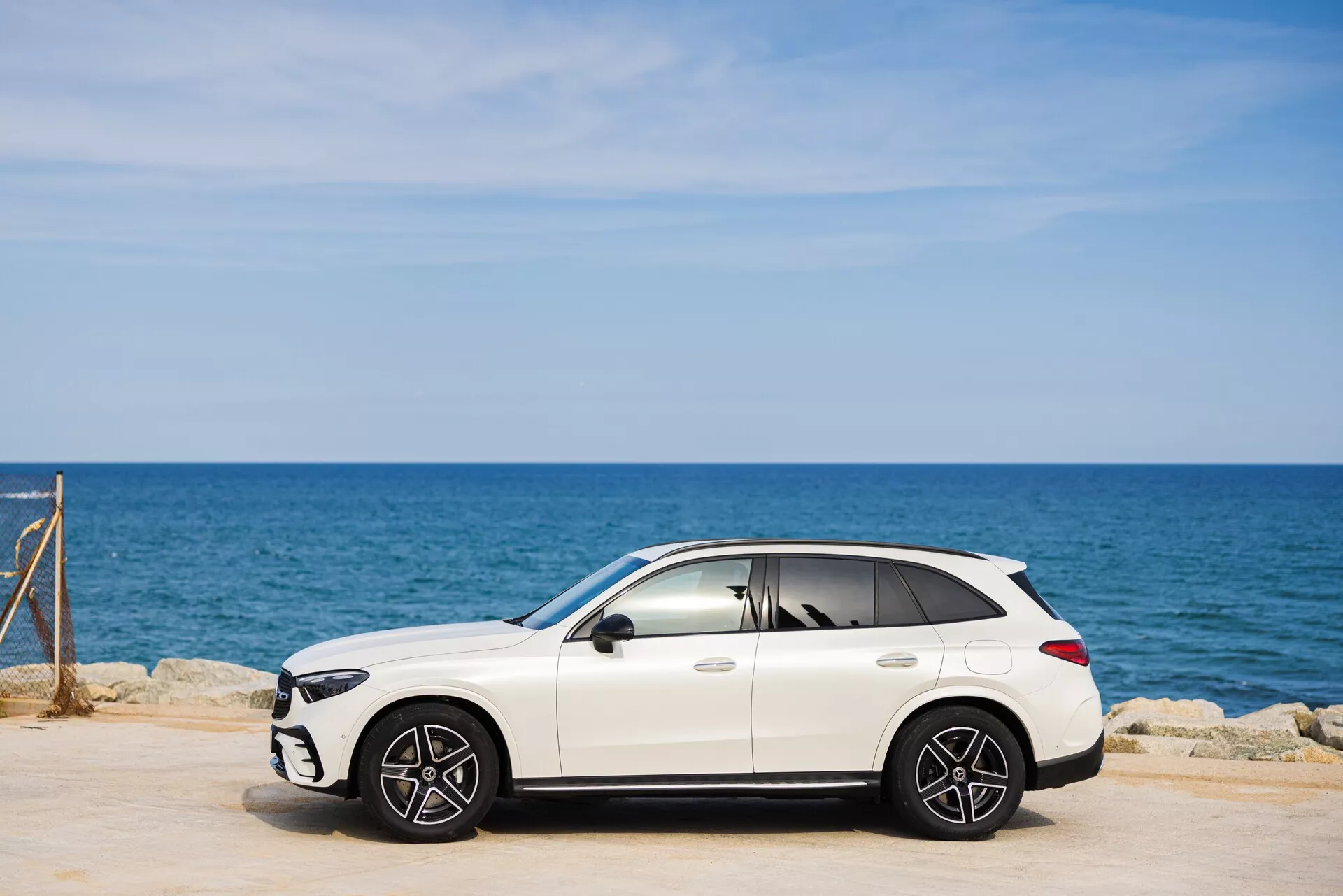 Slika belega luksuznega crossover SUV-ja Mercedes GLC 2022 (254), parkiranega na asfaltirani površini. Bočna perspektiva poudarja profil potniške strani z vidnimi sprednjimi in zadnjimi kolesi z več naperami. V ozadju so vidni morje in jasno modro nebo. Elegantna oblika SUV-ja vključuje stranska ogledala, kljuke na vratih in verjetno strešno okno. Sence pod vozilom kažejo na sončne pogoje. Rahlo zatemnjena okna dajejo občutek prestiža.