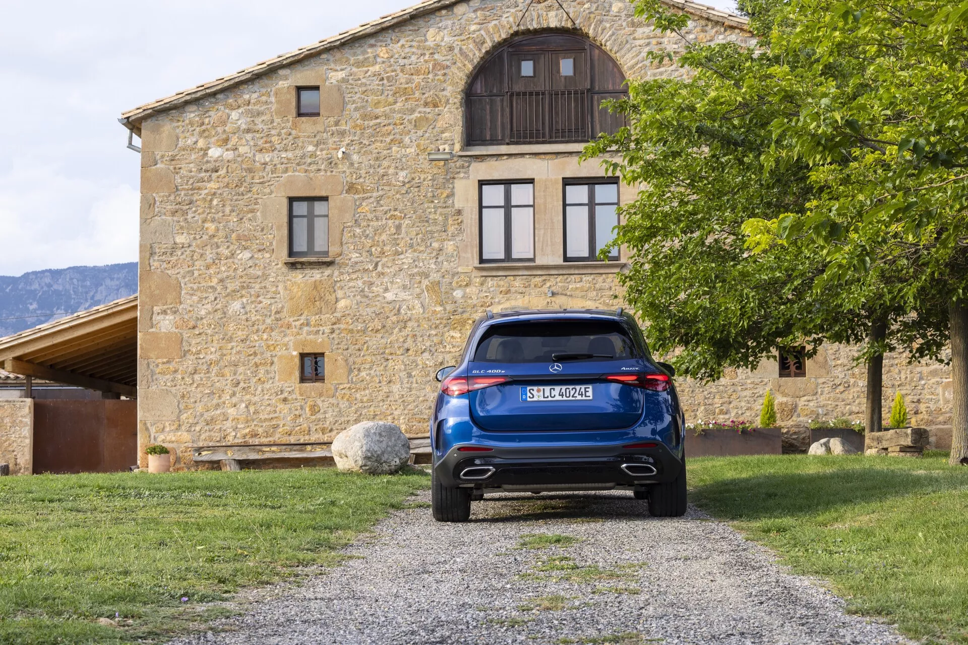 Zadnji pogled na moder Mercedes GLC 2022 (254), parkiran na gramozni poti, centriran v sestavi z zgradbo v ozadju. Slika poudarja prtljažna vrata, zadnje vetrobransko steklo z brisalcem, elegantne LED zadnje luči, zadnji odbijač s parkirnimi senzorji ali odsevniki in dvojni izpušni sistem. Značilnosti vključujejo visoko nameščeno zavorno luč, centralno nameščeno registrsko tablico, element difuzorja in strešni spojler, ki prikazujejo sodobno avtomobilsko tehnologijo in oblikovanje.