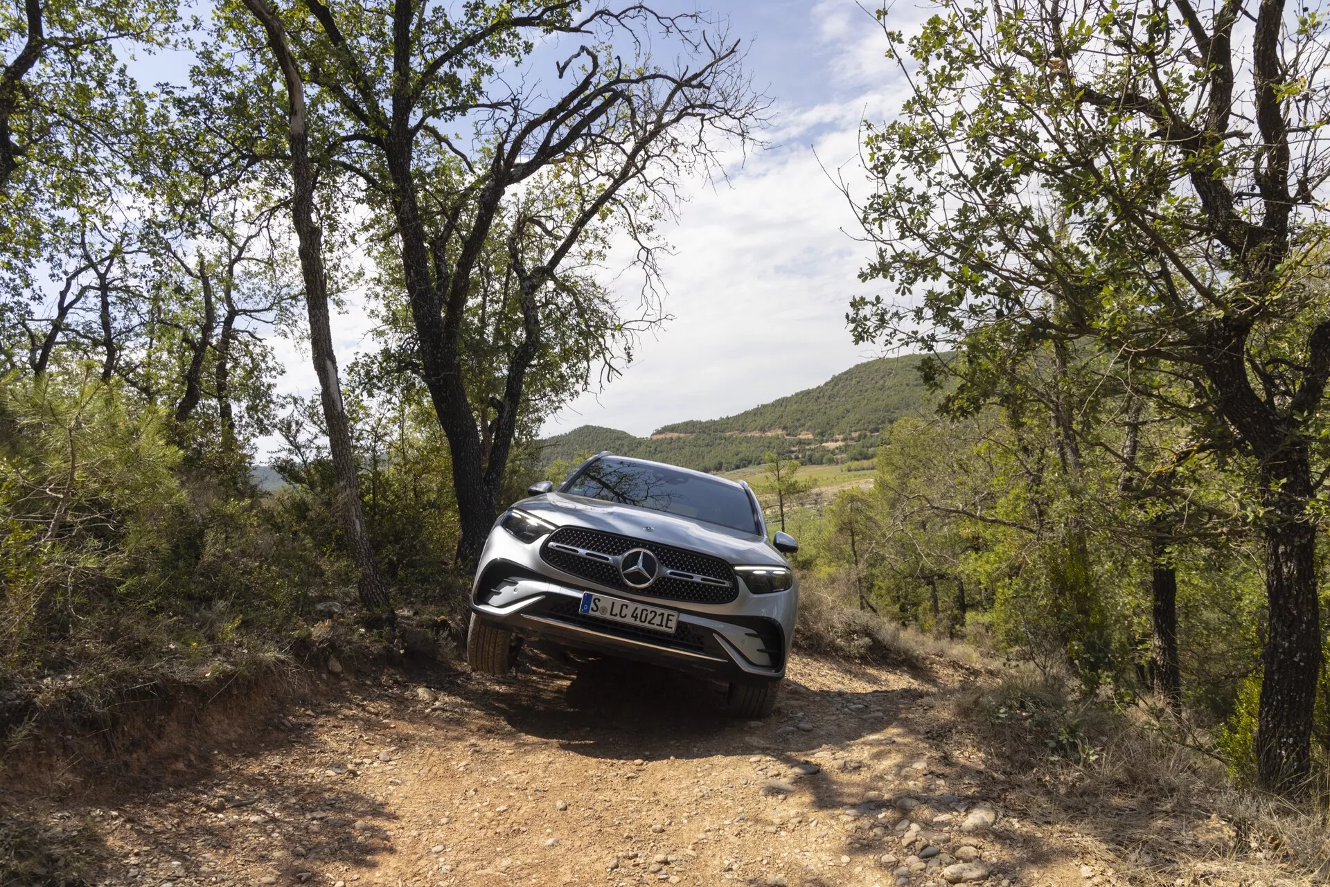 Barvna slika SUV Mercedes-Benz GLC 2022 v naravnem okolju, posneta iz nizkega sprednjega kota, ki poudarja njegovo robustno podobo. Fotografija se osredotoča na sprednjo fasado avtomobila, mrežo in moderne LED žaromete. Vozilo se pojavi na makadamski poti, obdano z zelenjem, kar poudarja njegove terenske zmogljivosti in eleganten dizajn. Sprednji senzorji in kamere nakazujejo napredne varnostne funkcije.