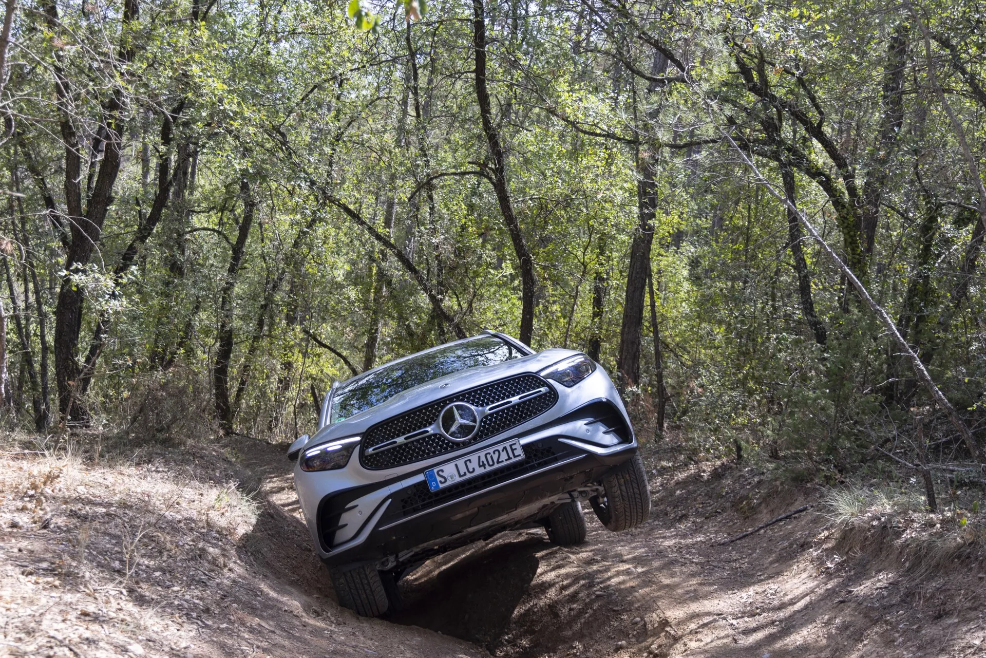 SUV Mercedes-Benz GLC 2022, ki se podaja po zahtevni, gozdnati off-road poti iz treh četrtin spredaj, prikazuje popolno rešetko, emblem, žaromete, del pokrova motorja in sprednji odbijač z zaščitno ploščo; stranska stran vidnejša z naklonjenim držanjem na neenakem terenu, obdana z drevesi in grmi, ki zajema off-road zmogljivosti SUV.