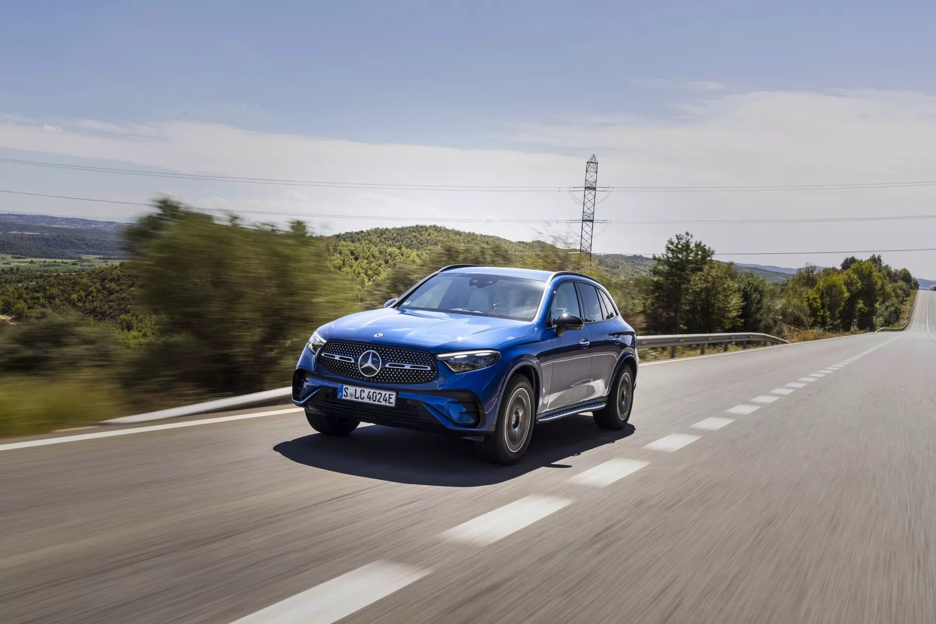Modri SUV Mercedes-Benz GLC 2022, ki se s hitrostjo vozi po podeželski cesti, posnet iz trikotne sprednje kote, prikazuje sprednjo masko, pokrov motorja, LED žaromete, ogledalo na voznikovi strani, kolesa in okna, z učinkom zameglitve gibanja na površini ceste in zelenje v ozadju, poudarja dinamično in luksuzno zasnovo avtomobila.