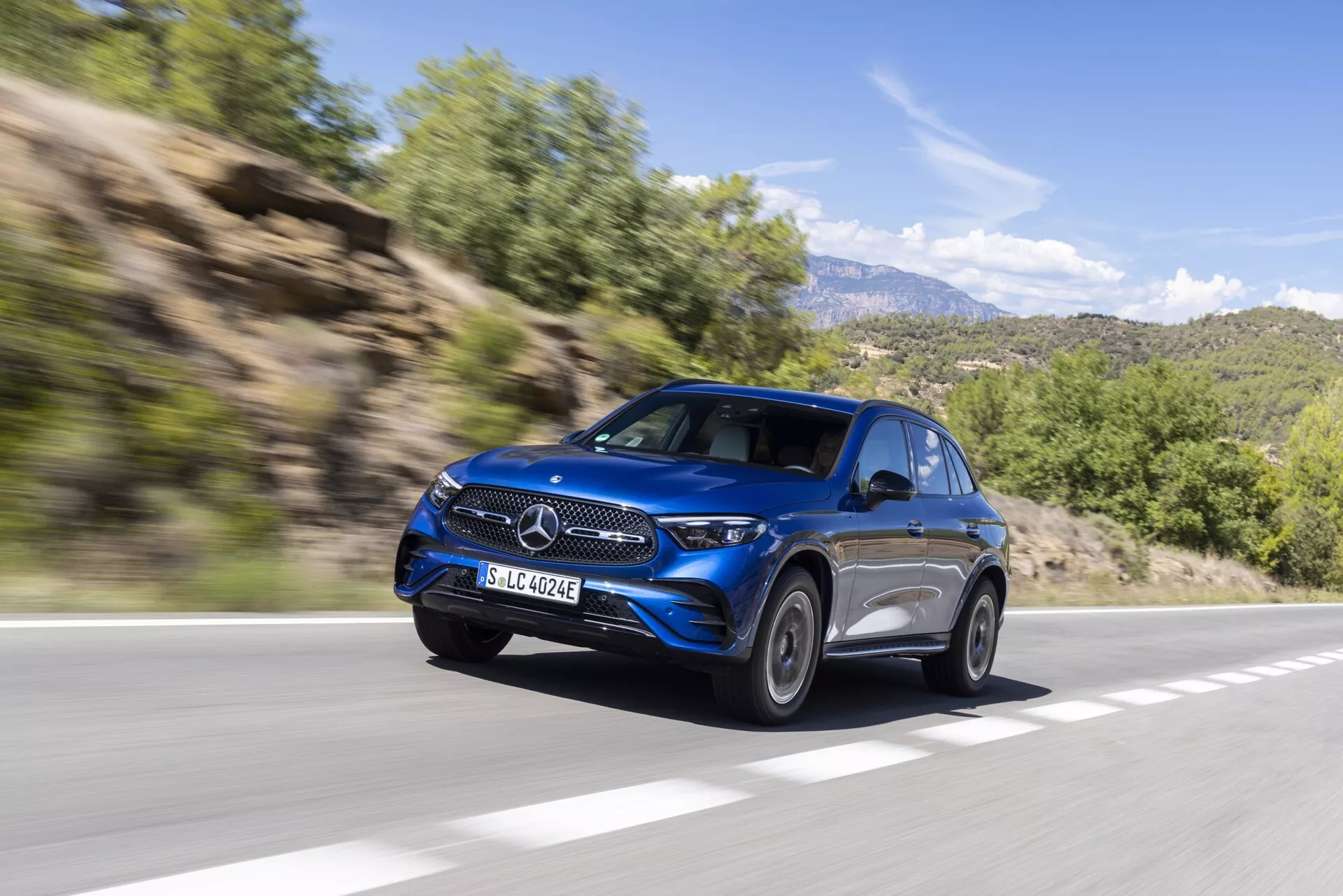Dinamičen posnetek modrega SUV-ja Mercedes GLC 2022 (254) v gibanju, videnega z levega sprednjega zornega kota. Sliko prikazujejo elegantne žaromete, izrazito rešetko z osrednjim emblemom, luksuzna lita platišča, aerodinamične linije karoserije in napredna stranska ogledala. Vozilo, ki vozi po asfaltirani cesti v podeželskem gorskem območju, ima zamegljeno ozadje z listjem in skalnatimi hribi, kar daje vtis hitrosti in sodobnega razkošja.