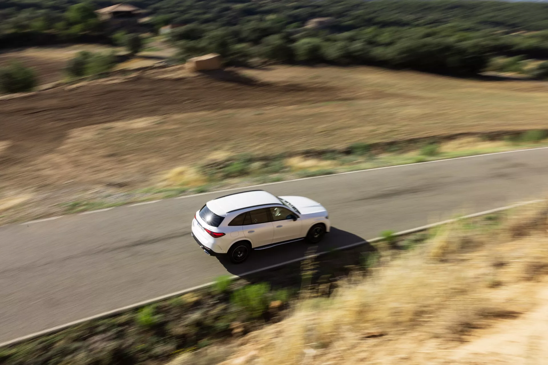 Visok kotni pogled na bele štirivratni SUV Mercedes GLC 2022 v gibanju na vijugasti podeželski cesti, obdani z naravno pokrajino. Vozilo je moderno aerodinamične oblike, s panoramsko streho, aluminijastimi platišči, zatemnjenimi stekli na zadnjih oknih in stranskimi ogledali z vgrajenimi smerniki. Zameglitev gibanja nakazuje hitrost in dinamično zmogljivost.