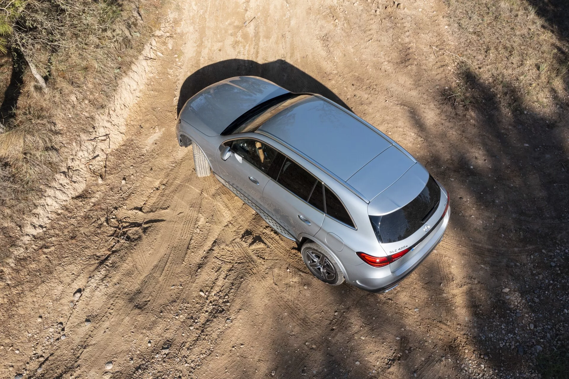 Pogled od zgoraj na srebrnega Mercedesa GLC 2022, ki prikazuje njegov sodoben dizajn z zaprtim strešnim oknom, zadnjo anteno in petkrakimi aluminijastimi platišči. Avto, postavljen na prašni podeželski cesti, poudarja njegovo možno obliko karoserije kot hatchback ali sportback, z sencami, ki nakazujejo jutranjo ali popoldansko sončno svetlobo.