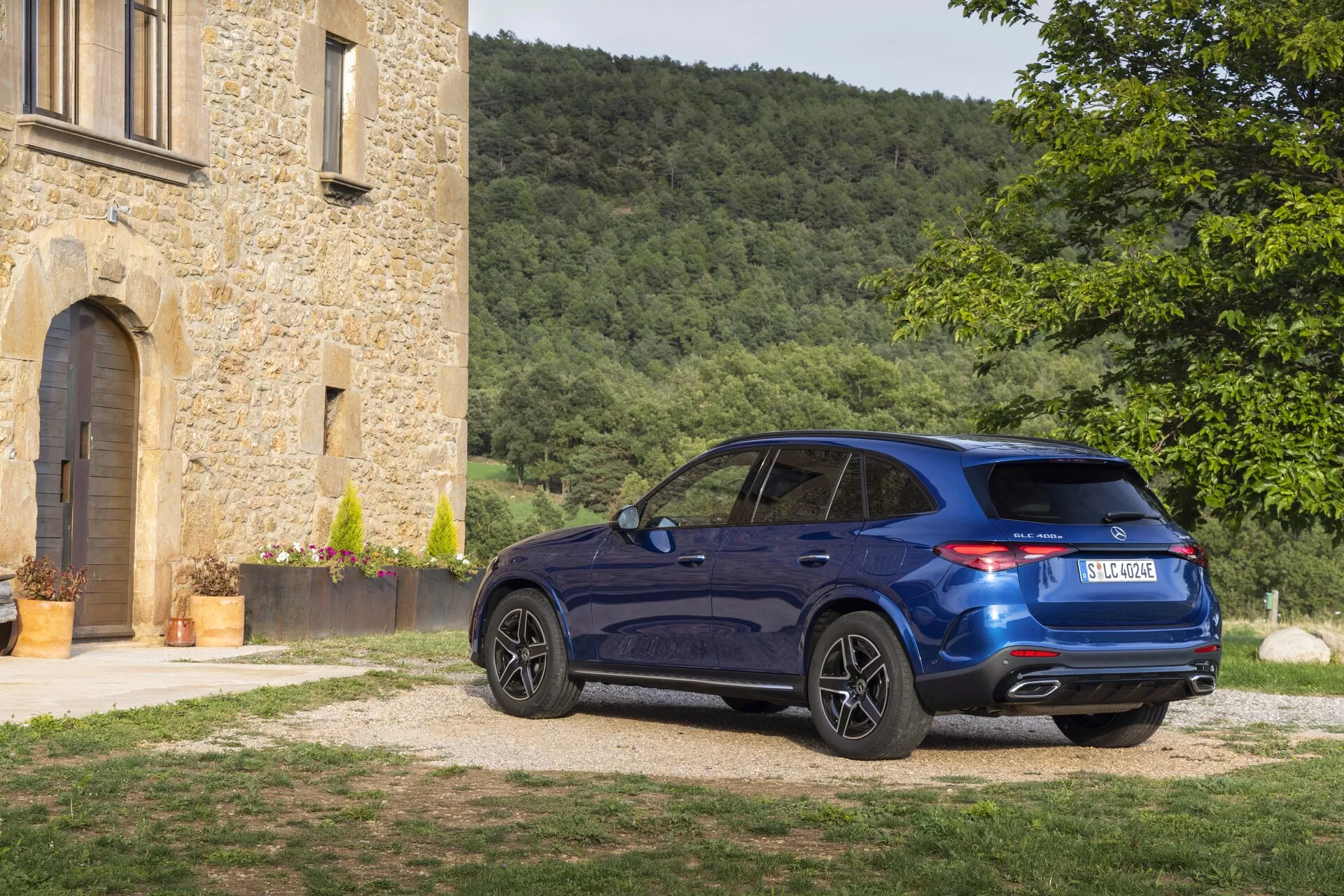 Luksuzni crossover SUV Mercedes-Benz GLC 2022 na gramozni dovozni poti poleg zgodovinske kamnite stavbe, iz tricatrtinske zadnje perspektive, prikazuje globoko modro barvo, LED zadnje luči, dvojni izpušni sistem, aluminijasta platišča in športno strešno linijo v opoldanskem osvetlitvi.