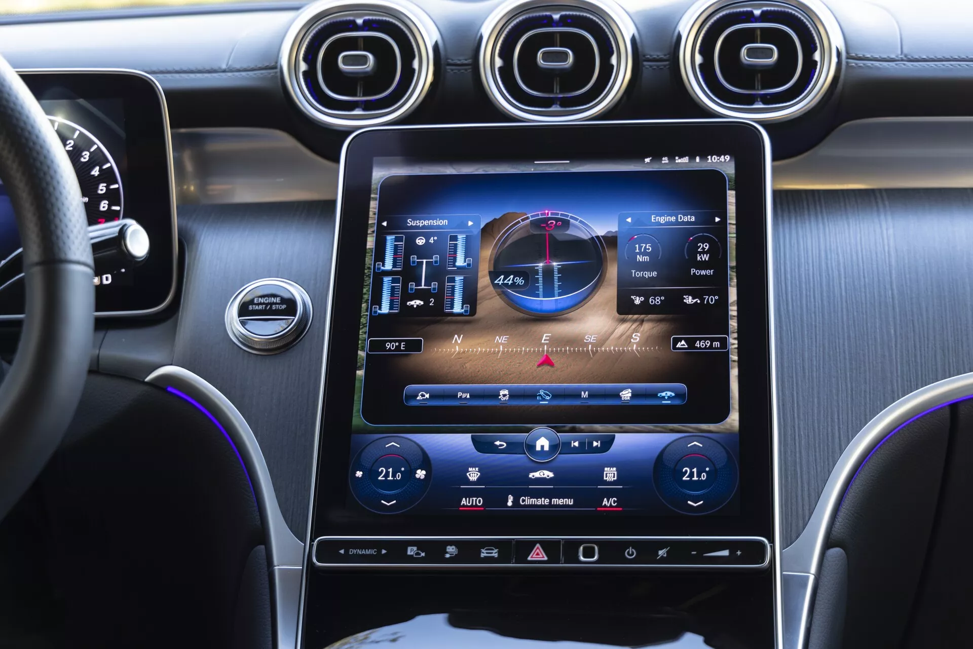 Pogled z višine oči s sedeža spredaj sodnik v notranjosti Mercedes GLC 2022, poudarja centralno konzolo z velikim navpičnim zaslonom na dotik z infozabavnim sistemom, ki prikazuje GPS, zvok in krmiljenje klimatskih naprav. Trije kovinski krožni zračniki nad zaslonom, namig armaturne plošče in volana na levi, vijolična ambientalna osvetlitev poudarja konzolo in armaturno ploščo.
