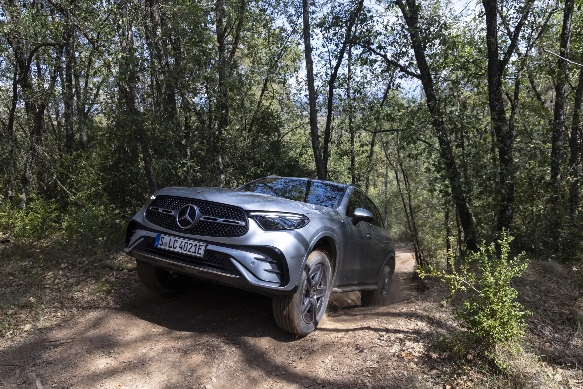 SUV Mercedes GLC 2022 postavljen na makadamski cesti obdani z gozdnim območjem, zajet iz sprednjega trikotnega kota, ki poudarja njegovo izrazito prednjo masko z emblemom znamke, LED žaromete, aluminijasta platišča in visoko oddaljenost od tal, poudarjajoč njegove terenske zmogljivosti v naravnem okolju dreves in suhega terena.