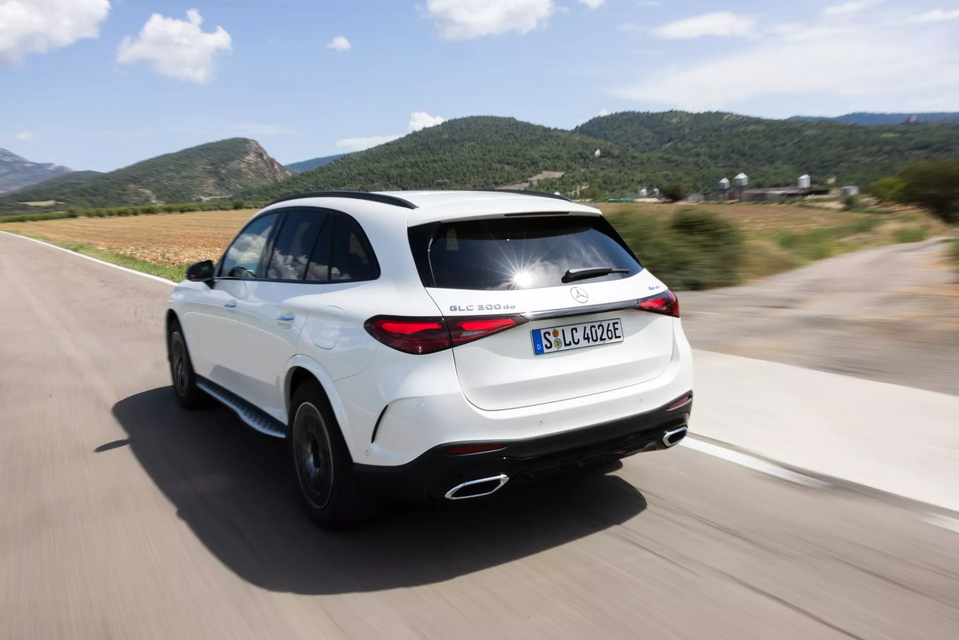 Dinamičen zadnji tritratinski pogled belega SUV-ja Mercedes GLC 254 iz leta 2022 v gibanju z učinkom panoramskega snemanja. Kot kamere zajame eleganten dizajn in moderna zadnja luči, prikaže desni zadnji kot, zadnje luči, zadnje kolo in del desnega stranskega profila na dobro vzdrževani cesti. Poudarja električno/hibridno različico z oznako 'EQ', športno držo, aerodinamične bočne krilce, dvojne izpušne cevi in strešno linijo v slogu kupeja. Zamegljeno ozadje in kolesa poudarjajo hitrost in gibanje avtomobila.