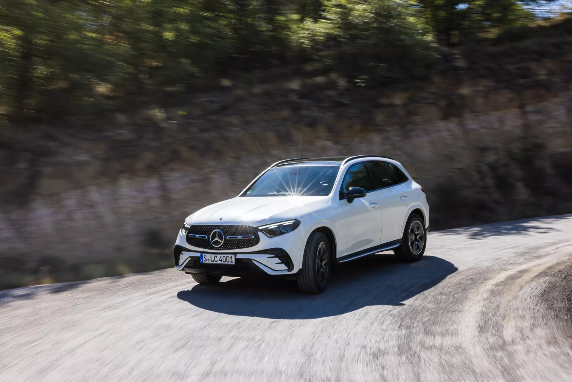 Dinamičen posnetek belega Mercedes-Benz GLC 2022 SUV v gibanju, posnet podnevi z treh četrtin spredaj. Poudarjeni so sprednja mreža z izrazitim emblemom, značilne LED luči, sprednji odbijač z vidnimi odprtinami za zrak, oblikovana kapuca z dvignjenimi linijami in stranski profil do zadnjih vrat. Kolesa so prikazana v gibanju z rahlim zamegljevanjem, kar nakazuje hitrost. Stranska ogledala so aerodinamično oblikovana. Okolje se zaradi gibanja vozila zdi zamegljeno, kar poudarja dinamično in hitro naravo avtomobila.